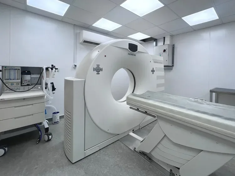 A medical imaging room with a Siemens MRI machine, a nearby control cart, and medical equipment, all in a clean, white-walled space with bright ceiling lights.