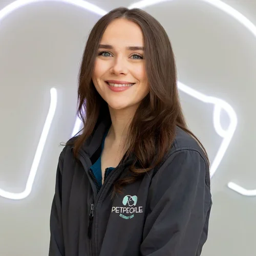 A smiling woman with long brown hair wearing a black jacket with a 'PETPEOPLE Veterinary Care' logo standing in front of a white wall with neon lights.