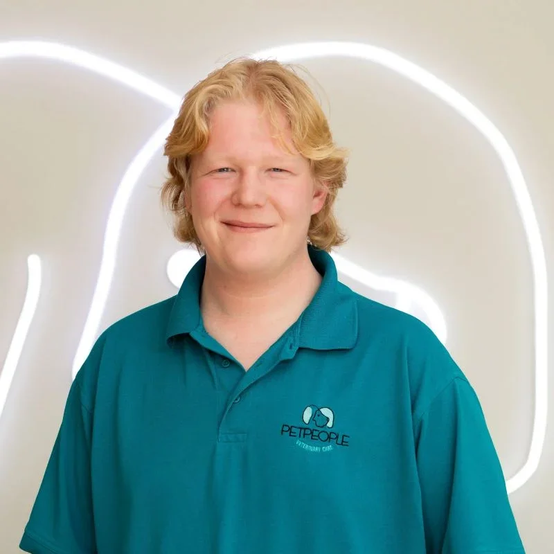 A young man with blond hair wearing a teal polo shirt with a pet care logo. He is standing in front of a white wall with a glowing pet silhouette outline.