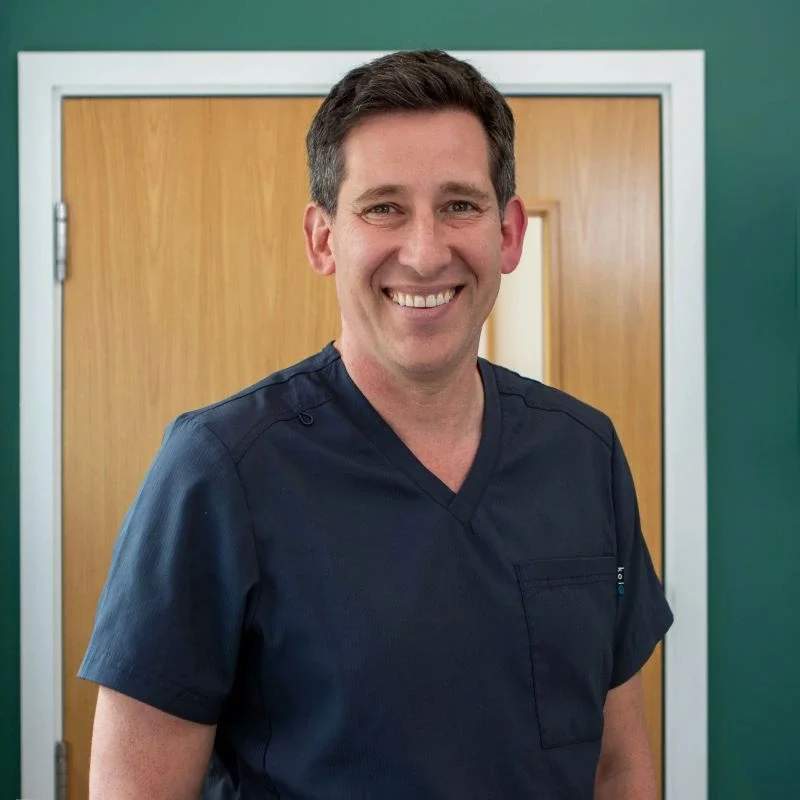 A smiling man in navy medical scrubs standing in front of a wooden door.