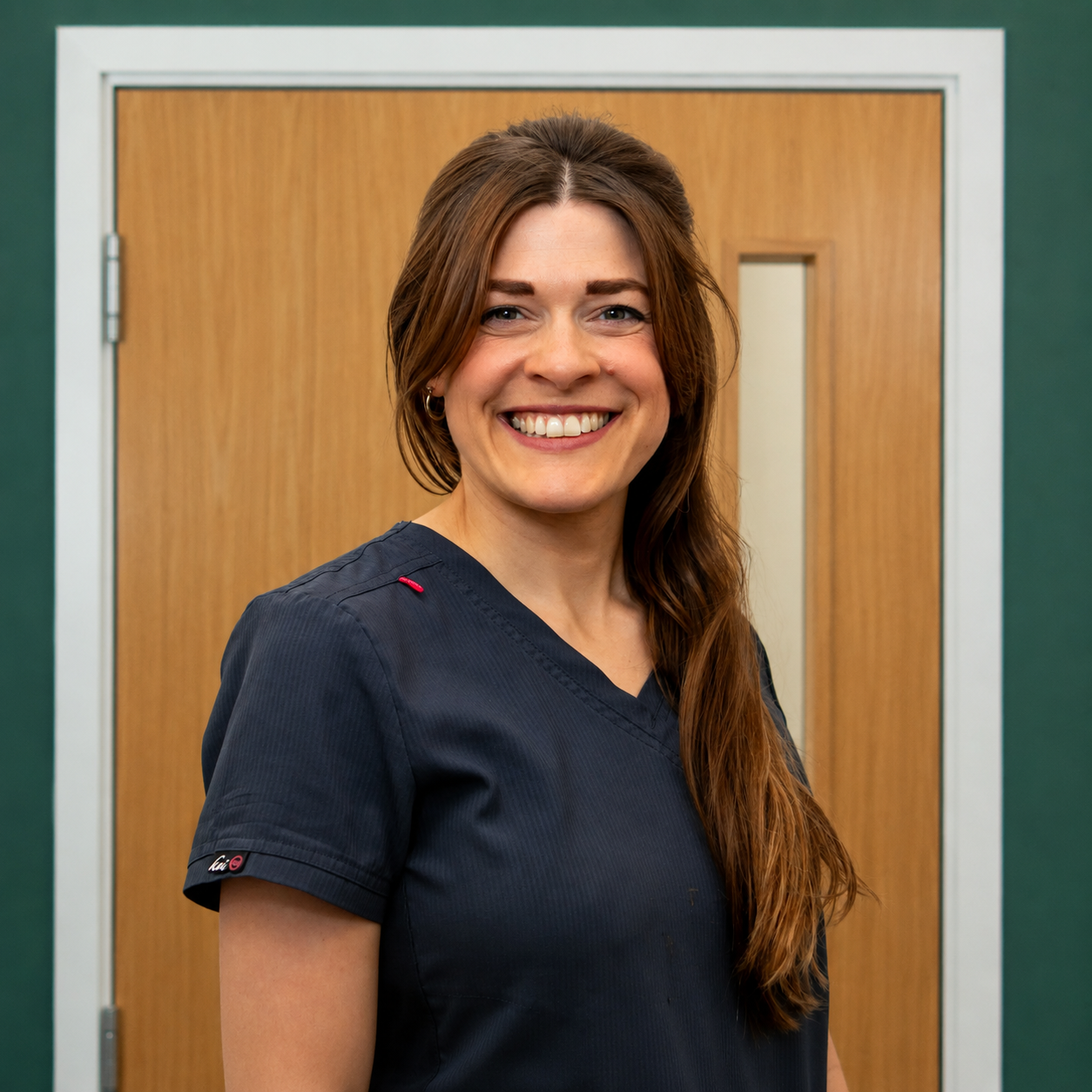 A smiling woman with long brown hair wearing navy medical scrubs standing against a plain white background.