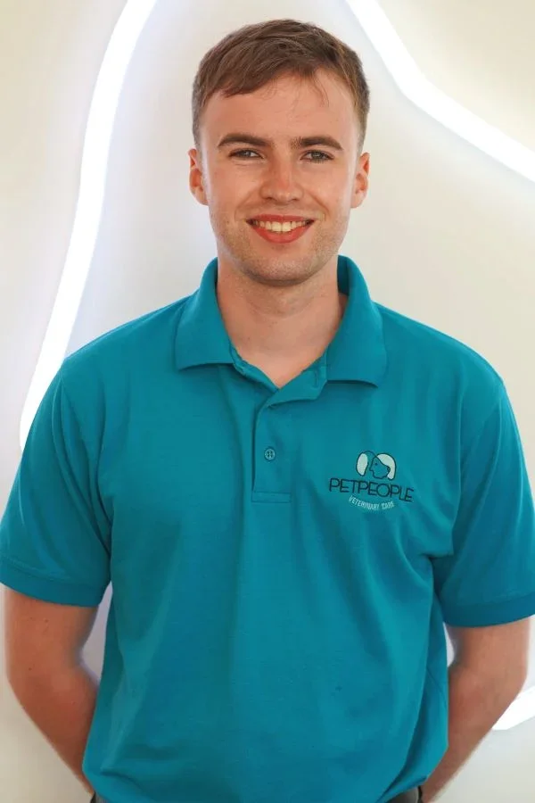 A young man stands in front of a background with abstract white and beige shapes. He is wearing a teal polo shirt with a pet-themed veterinary logo on it.
