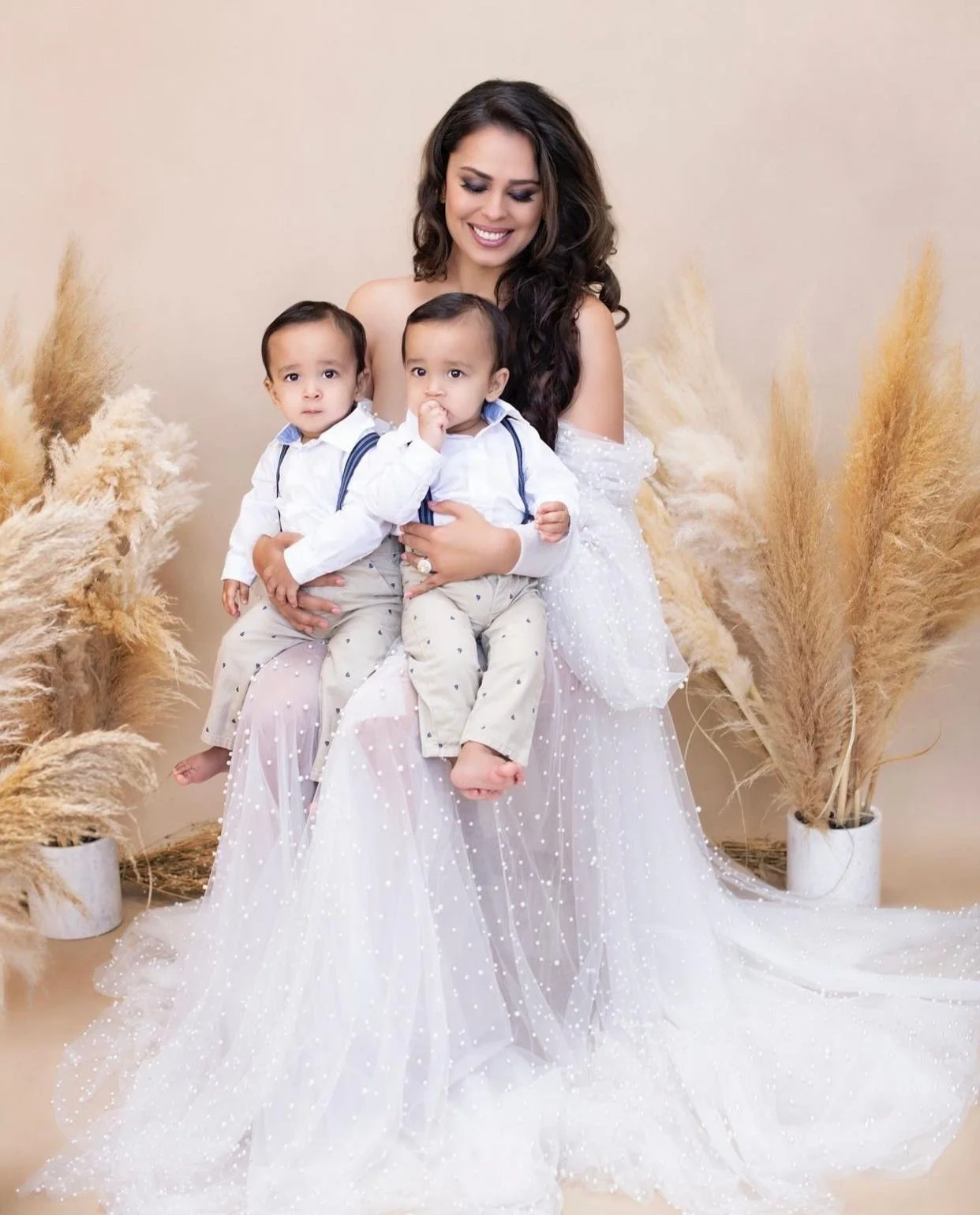 A woman in a white, sparkly dress sitting on a chair, holding two young boys on her lap. The boys are wearing white shirts with suspenders and beige pants. The background has beige walls and two vases with dried pampas grass.