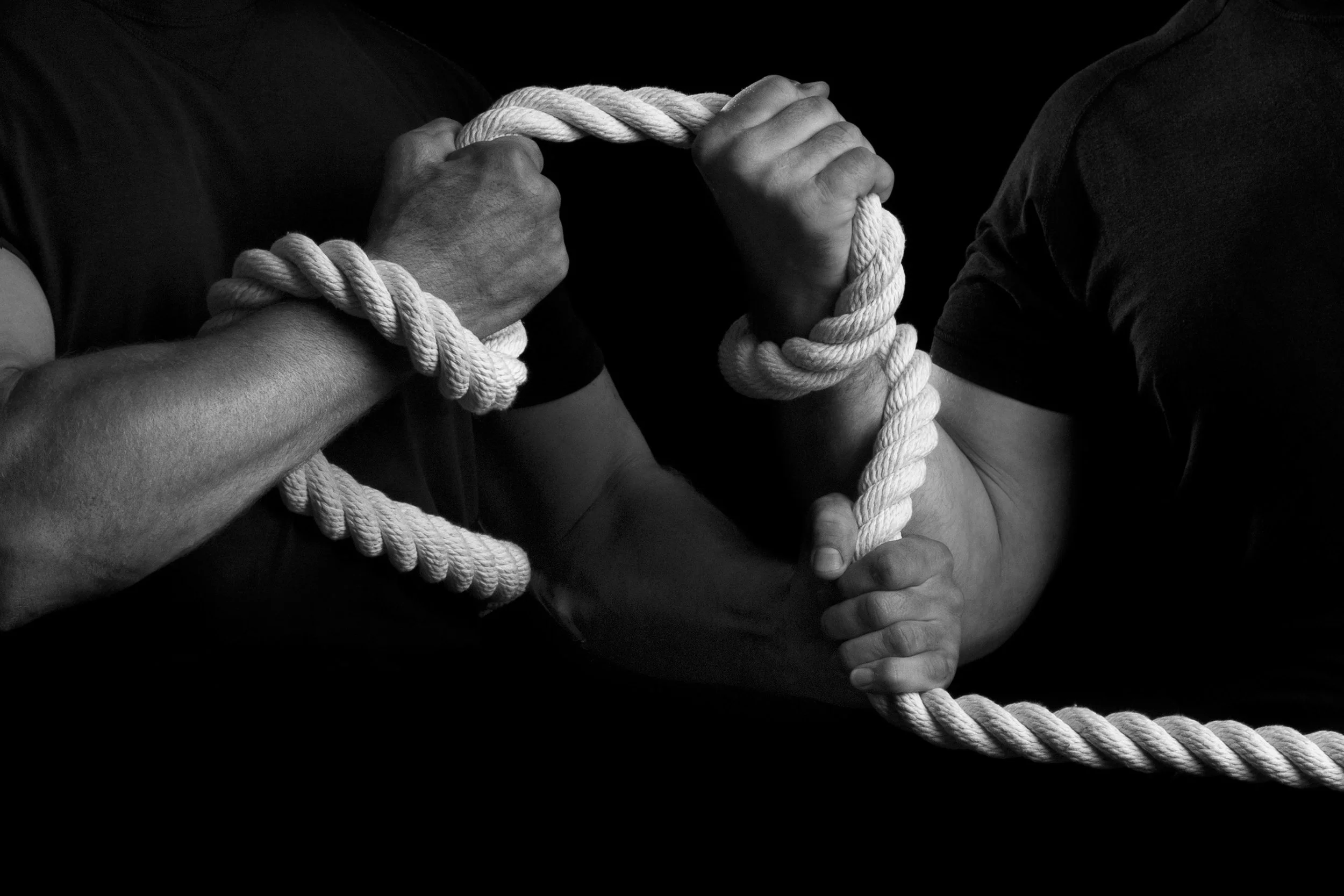 Two people in black shirts wrestling with a thick, white rope, each gripping it tightly with both hands.