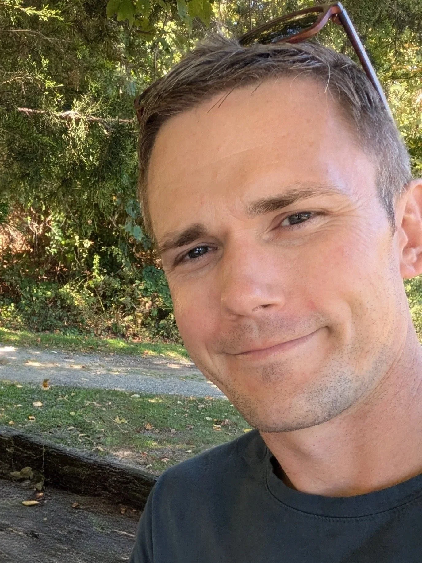 Close-up of a young man with short brown hair, light skin, wearing a dark shirt, and sunglasses resting on his head, smiling outdoors with trees and greenery in the background.