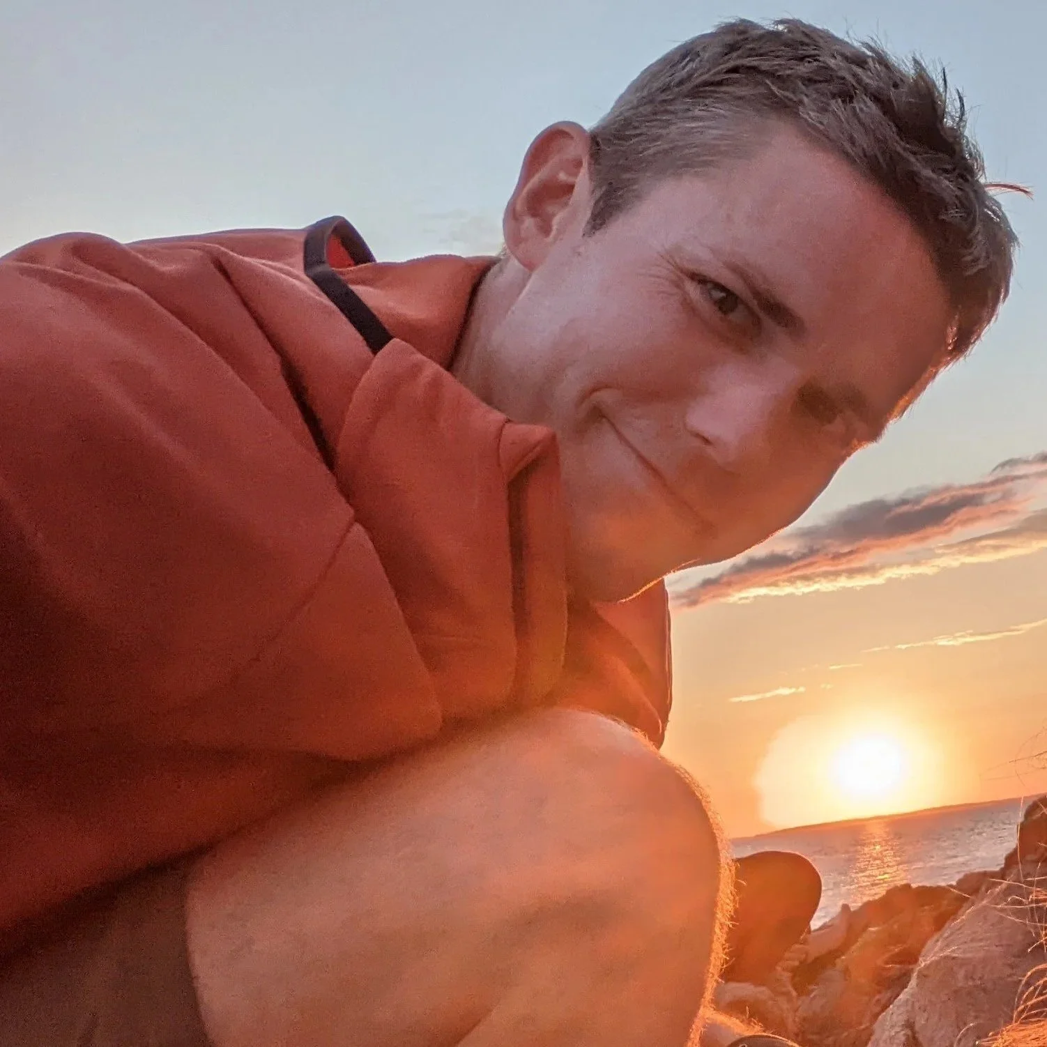 A smiling man with short hair wearing a reddish jacket, sitting near the ocean rocks during sunset, with the sun setting over the water in the background.