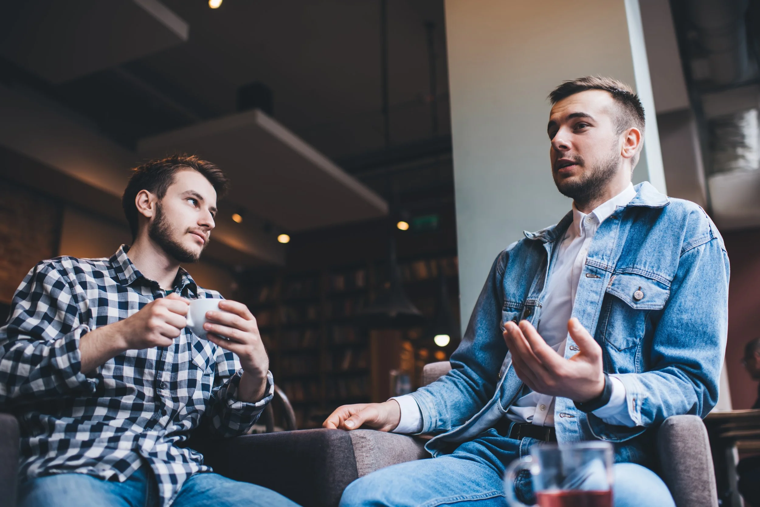 Two young men having a conversation in a coffee shop, with one holding a coffee cup and the other gesturing with his hand.