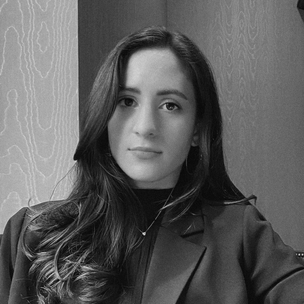 Black and white portrait of a young woman with long hair, wearing a blazer and hoop earrings, looking at the camera.