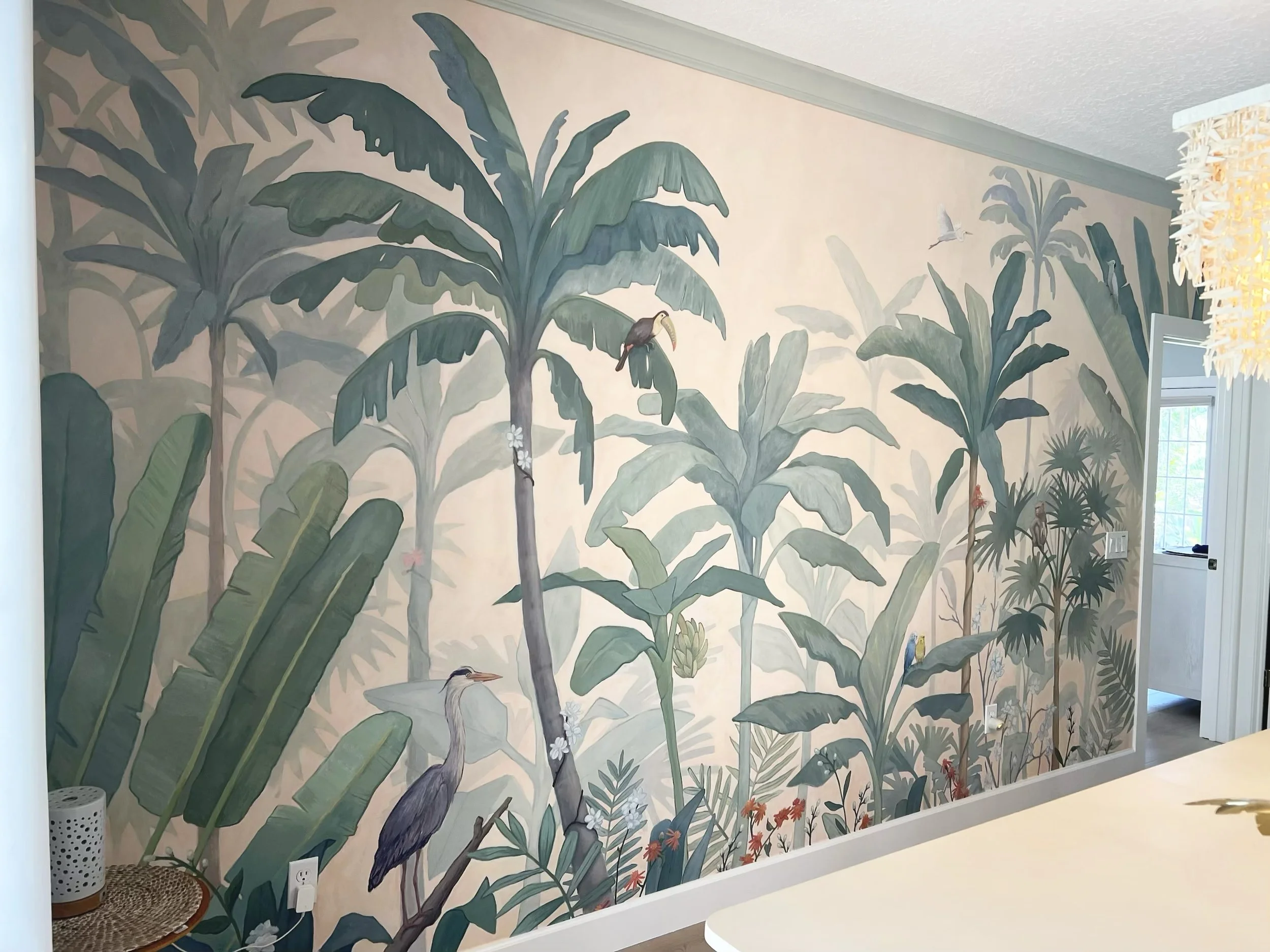 A wall mural depicting a lush jungle scene with tall green banana leaves, various tropical plants, birds including a toucan and heron, and a hummingbird, all painted in a soft, pastel color palette.
