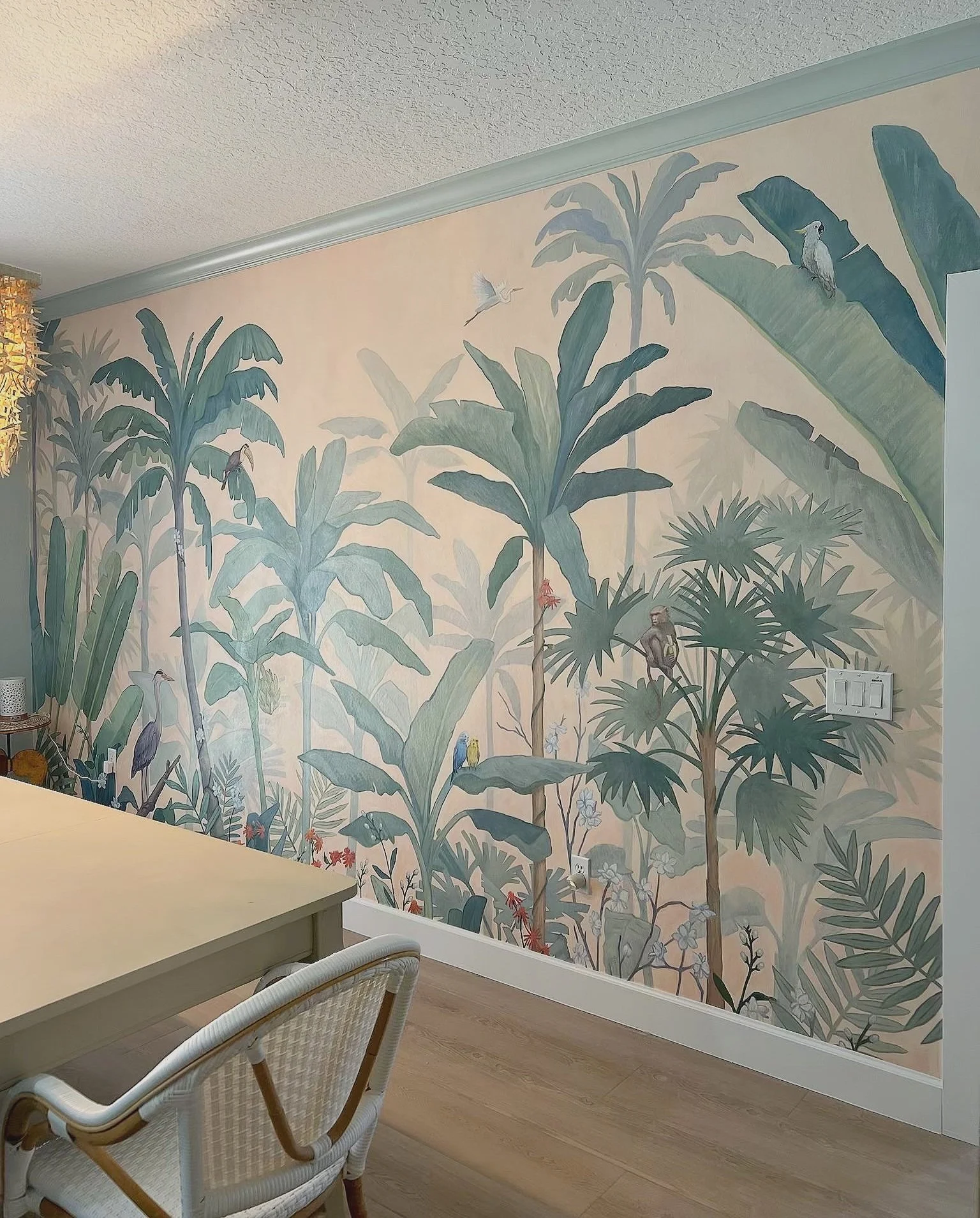 A tropical forest mural on a wall featuring large green leaves, palm trees, parrots, toucans, and other tropical birds in a colorful landscape, with a table and chairs in the foreground.