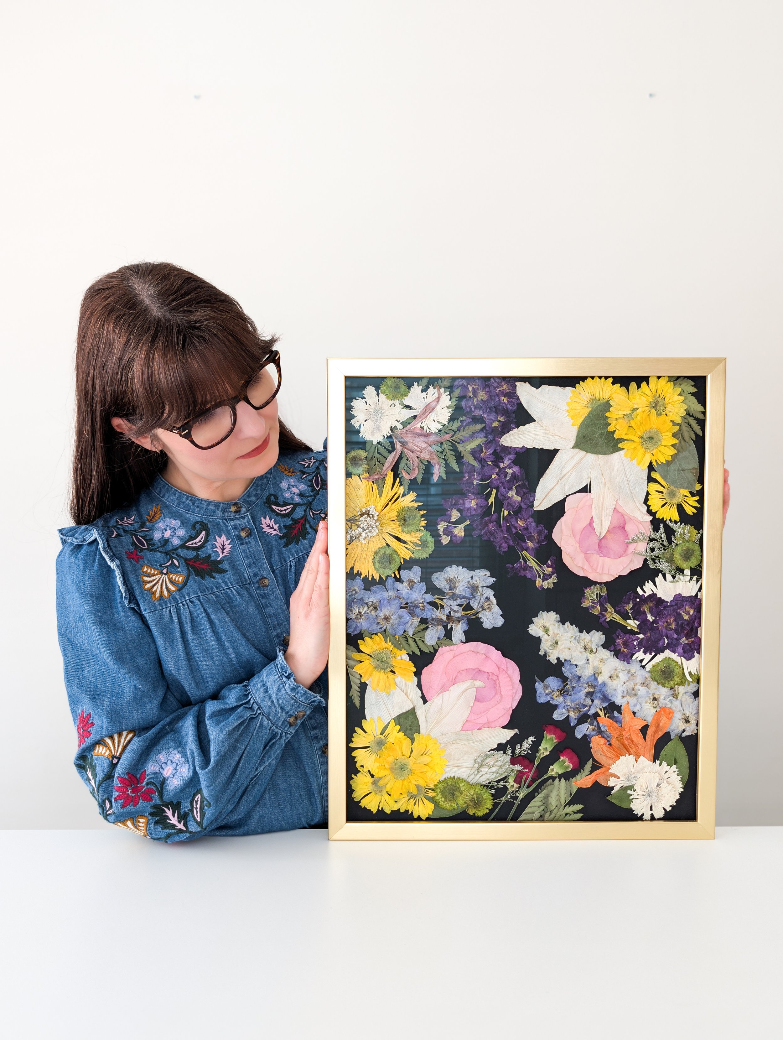 Woman with glasses in a denim embroidered shirt holding a floral framed artwork featuring various colorful flowers against a black background.