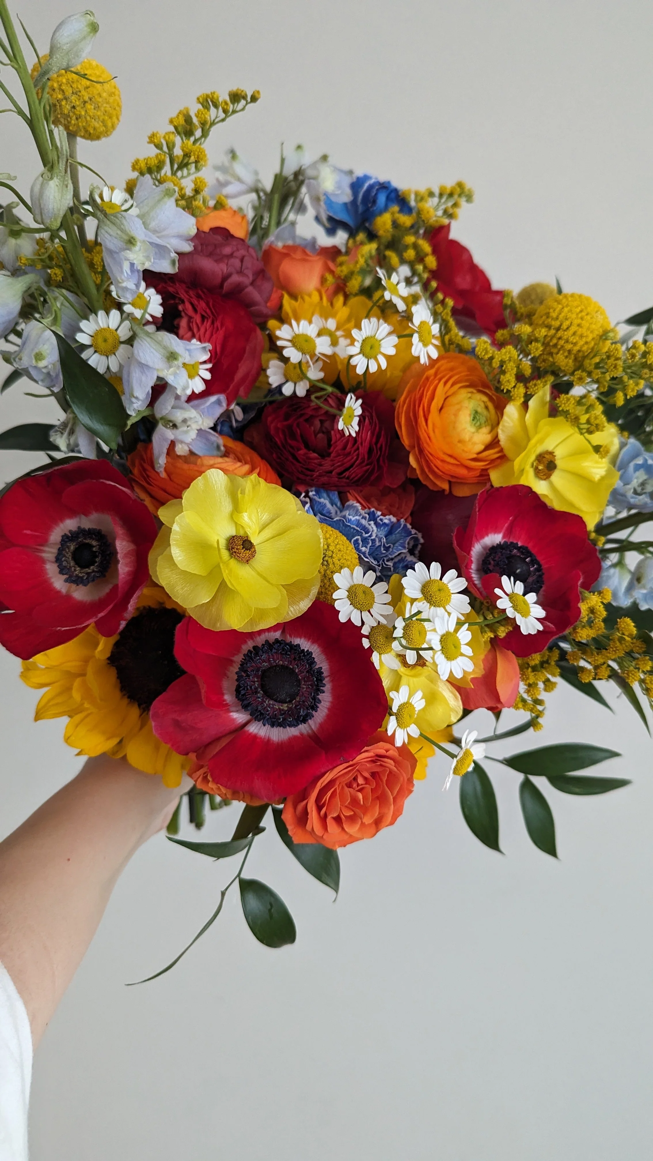 A colorful bouquet of mixed flowers including red poppies, yellow ranunculus, white daisies, yellow billy buttons, purple irises, blue flowers, and greenery against a plain white background.