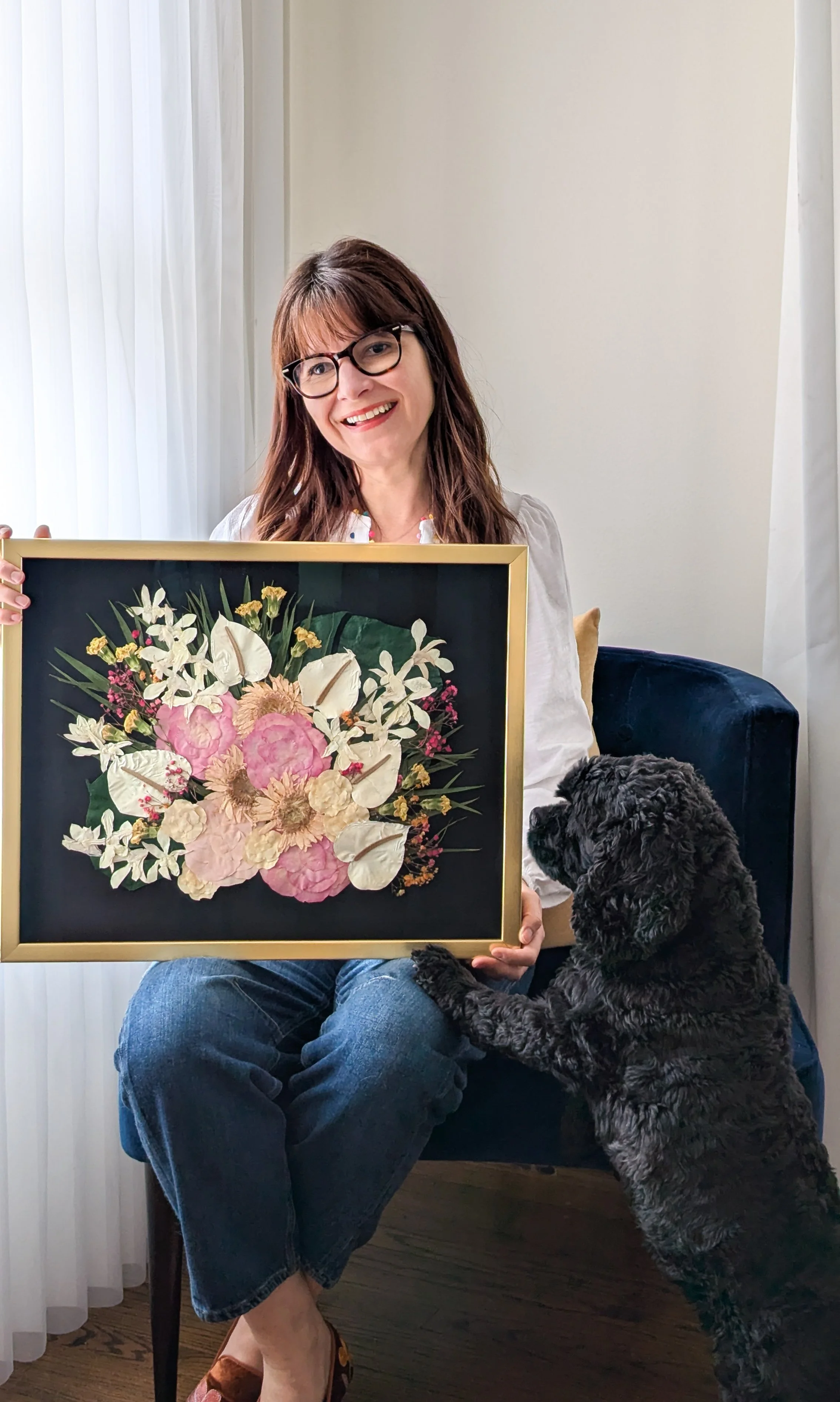 A woman with glasses and brown hair sitting on a chair, holding a floral artwork, while a black curly-haired dog touches the artwork with its paw.