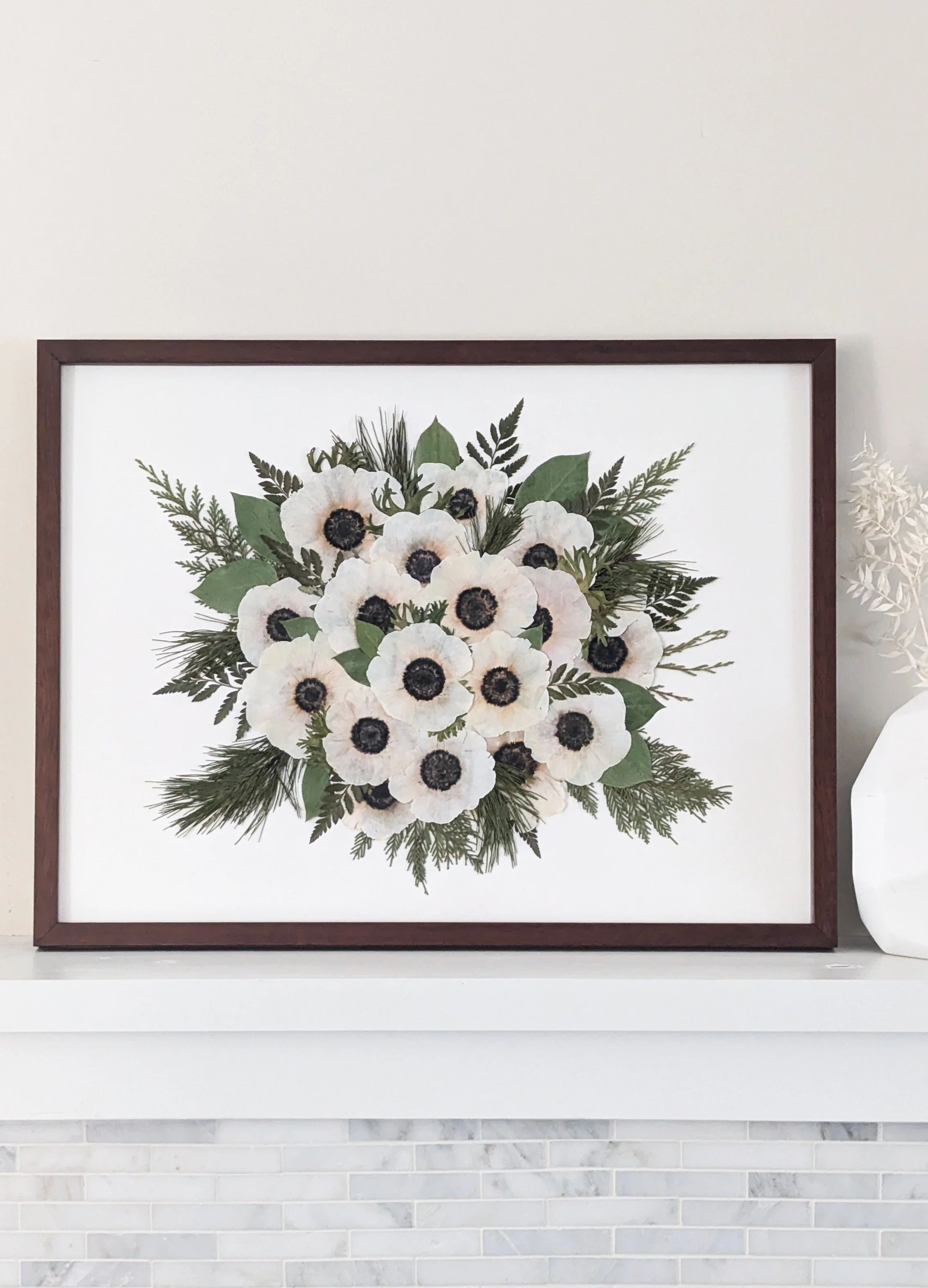 A framed artwork of a bouquet of pressed white anemone flowers with black centers and green leaves, placed on a white surface next to a white decorative object on the right.