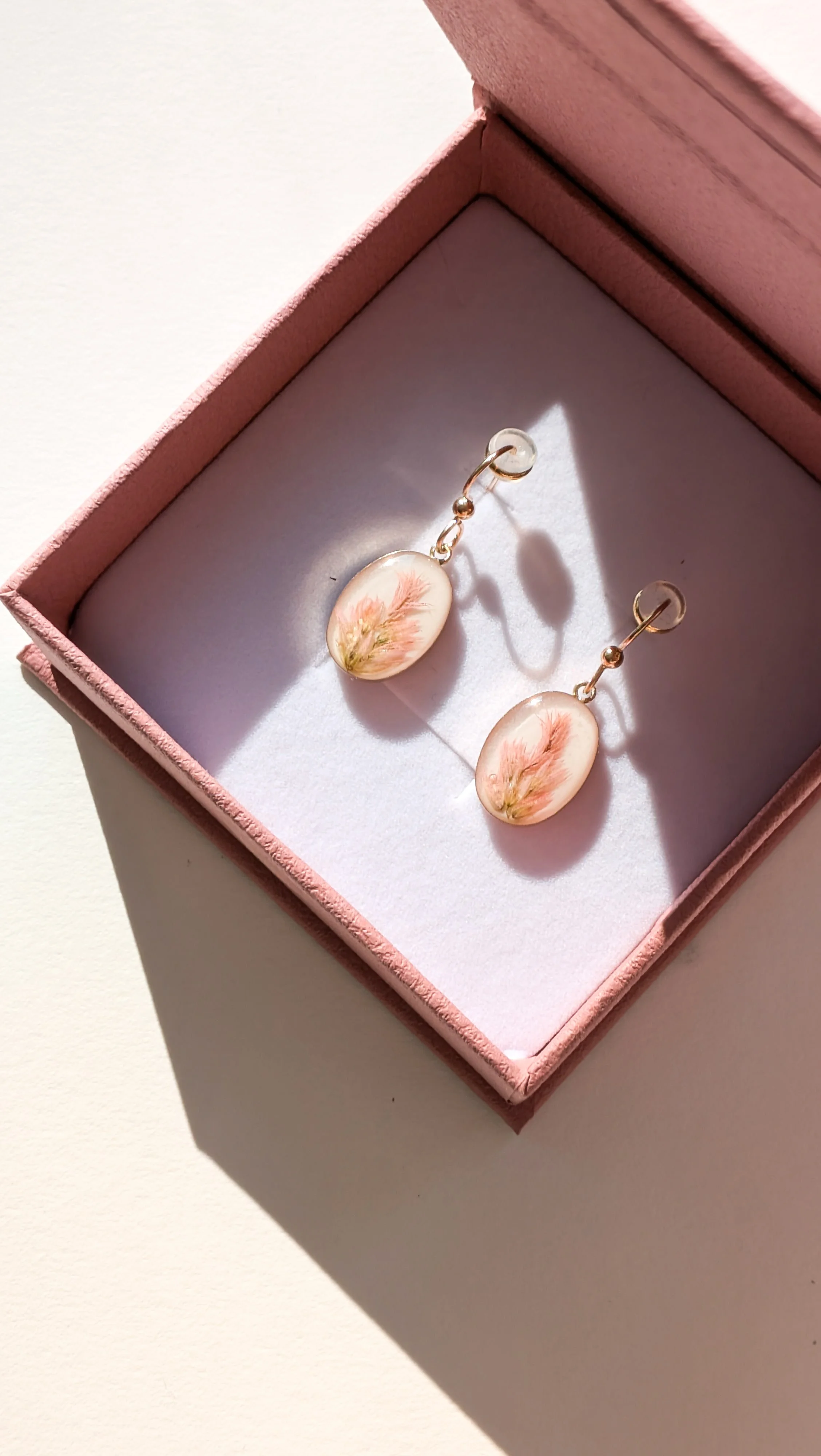 A pair of earrings with pink floral designs inside an open pink jewelry box on a white surface.