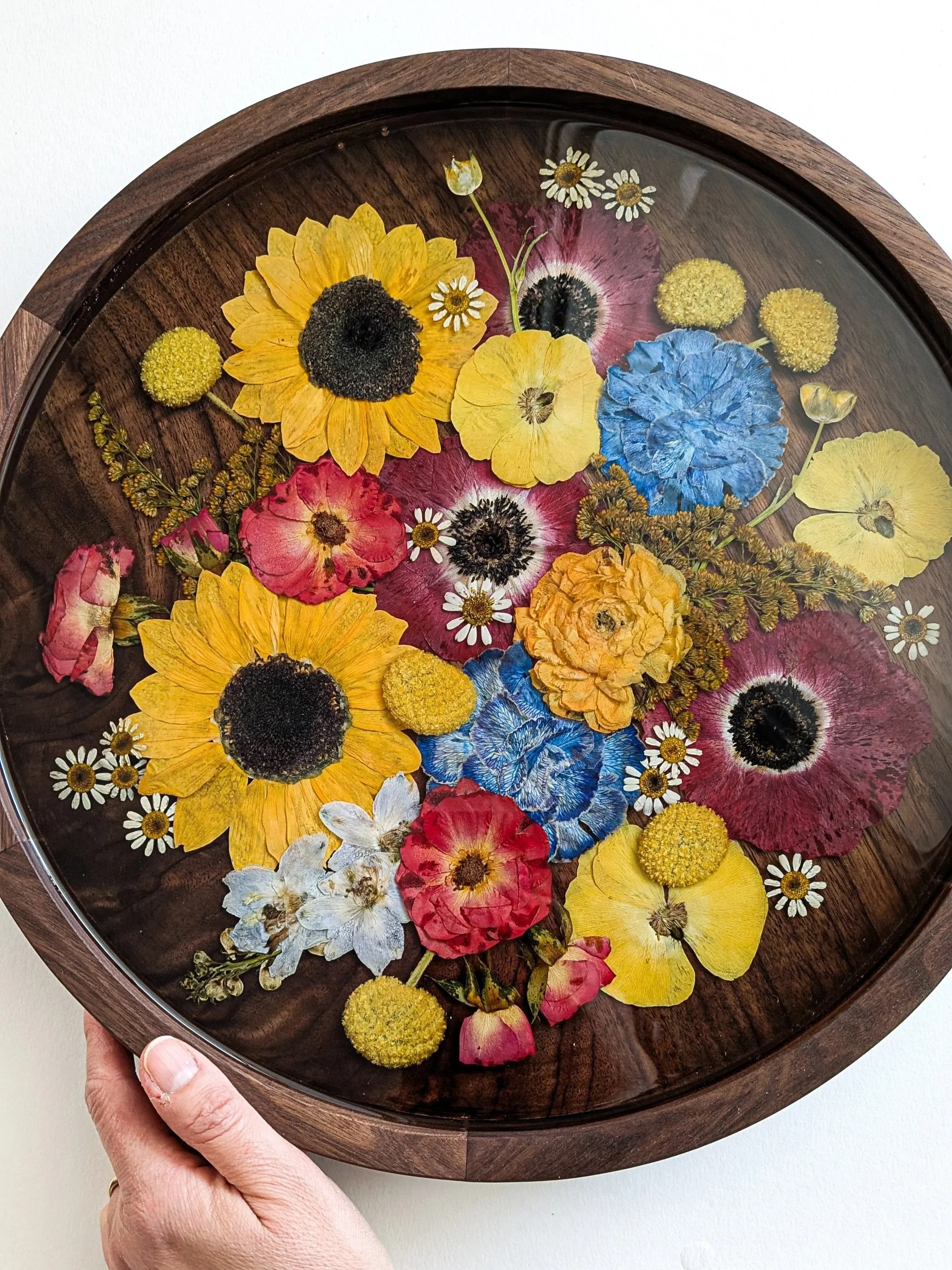 A round wooden tray filled with colorful pressed flowers under a glass cover, including sunflowers, daisies, and other various blossoms.