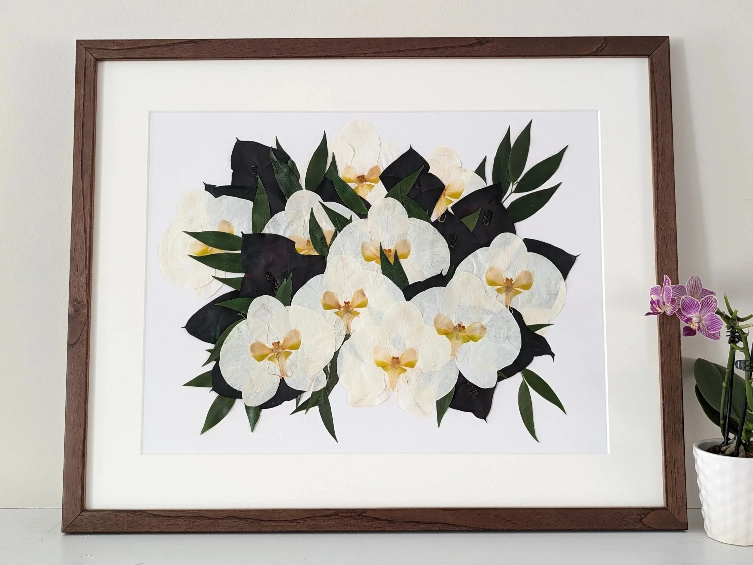 Framed botanical artwork of white and black orchids with green leaves on a white background, placed on a light-colored surface, next to a white pot with a pink orchid.