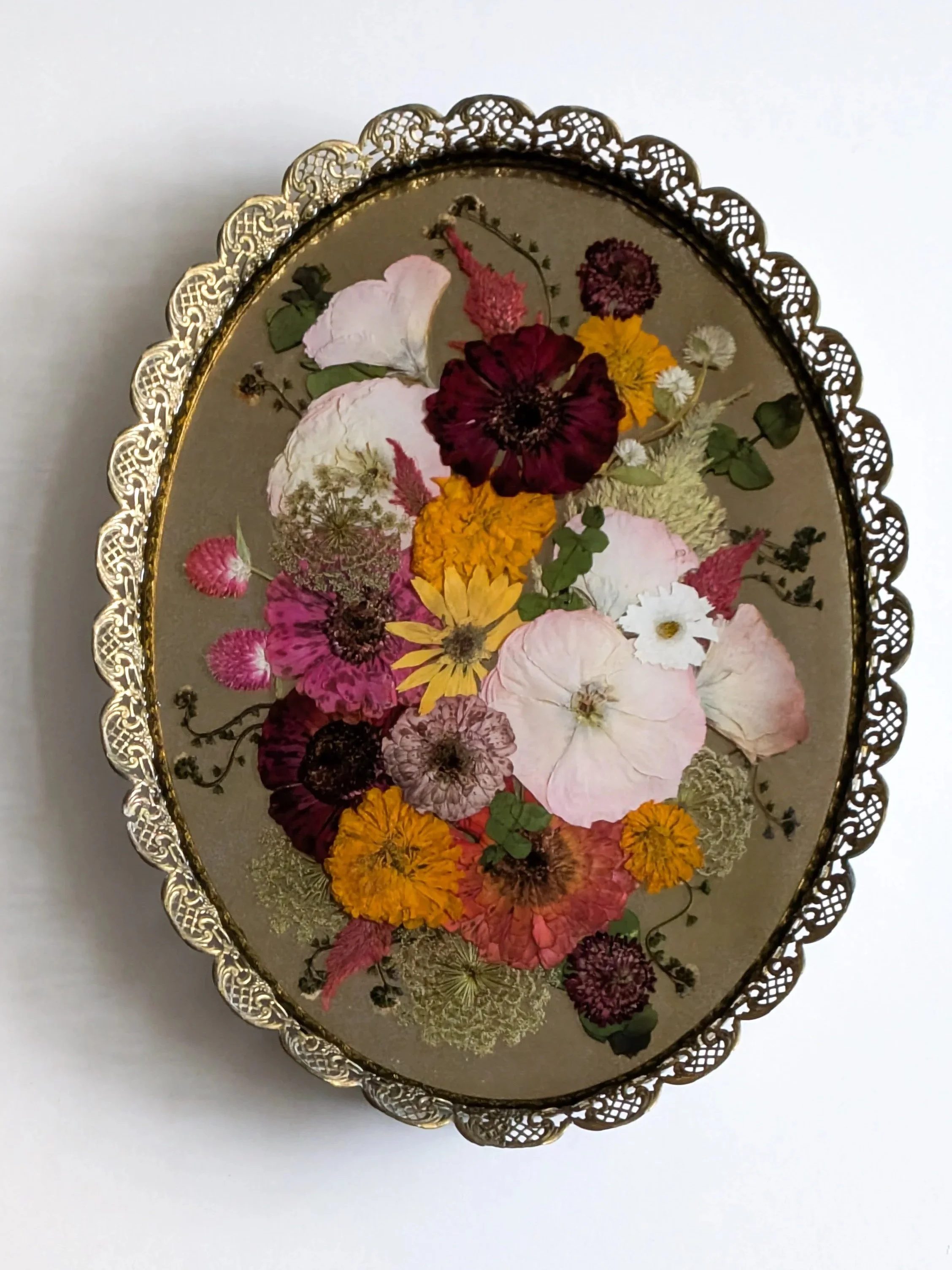 Oval glass art piece with dried pressed flowers and leaves, framed with ornate gold metal edging