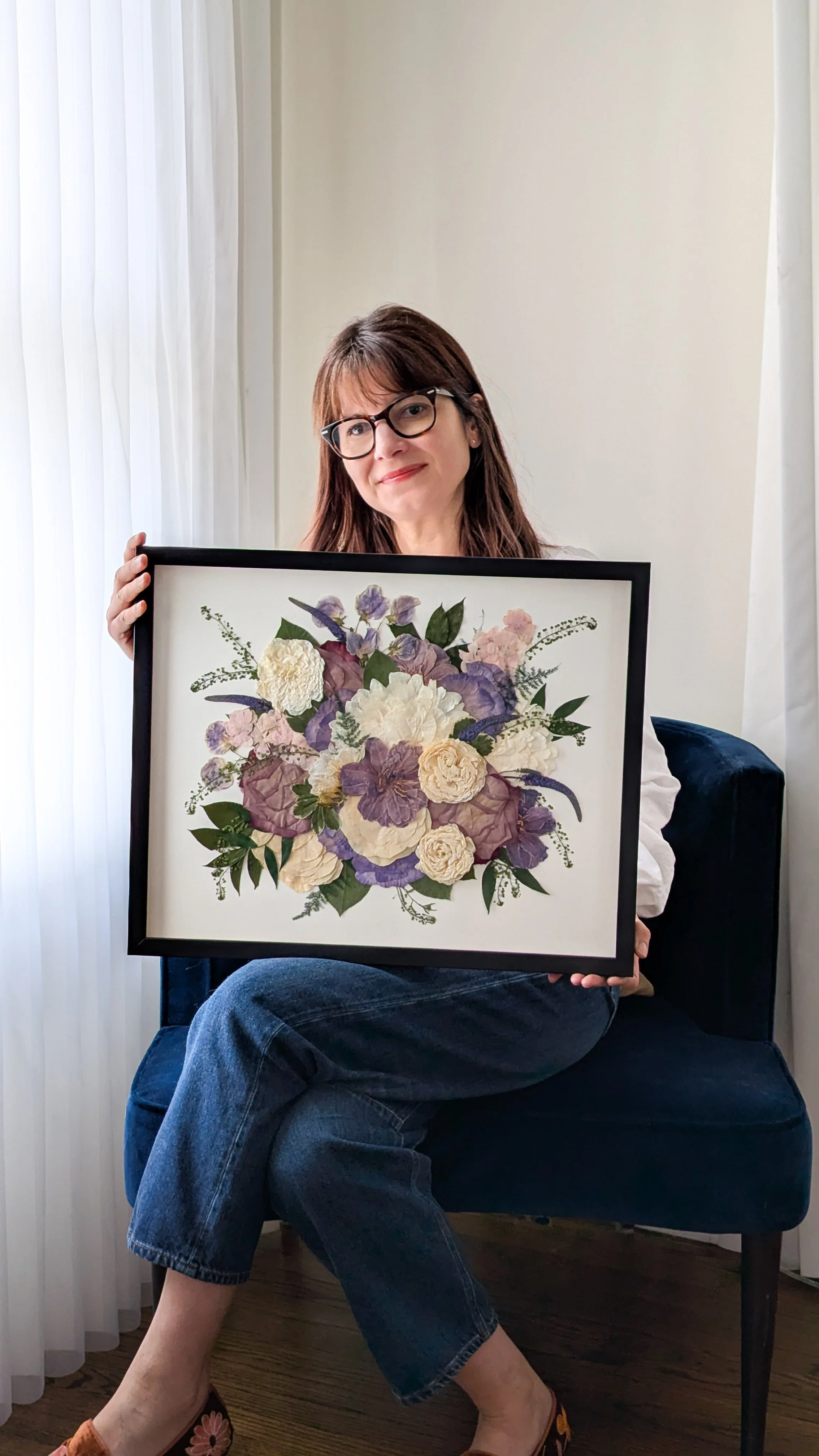 Woman sitting on a blue chair holding a framed floral artwork with white, purple, and pink flowers and green leaves, near a window with white curtains.