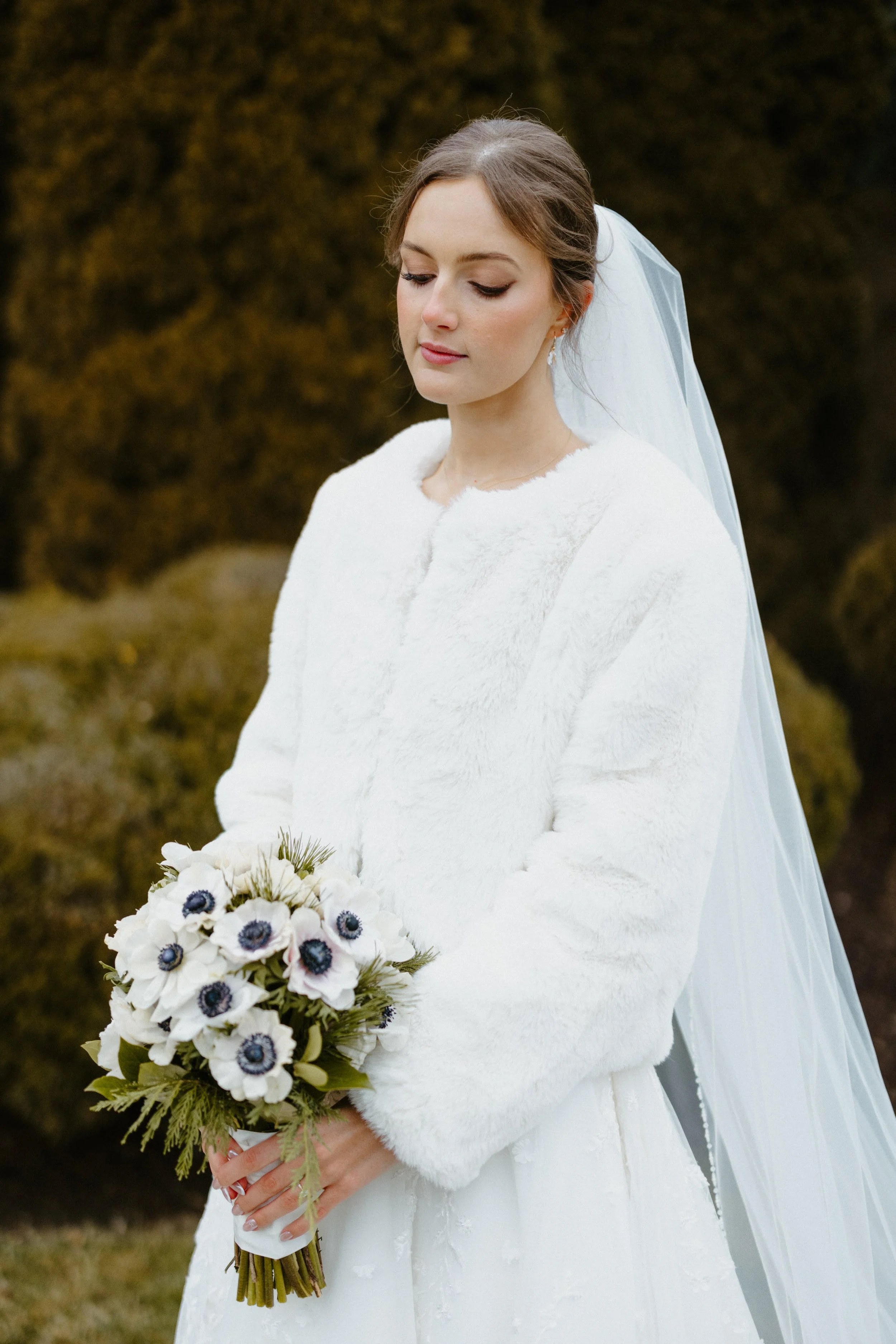 Bridal portrait of a woman in a white wedding dress with veil, holding a bouquet of white anemone flowers with dark centers, standing outdoors with greenery background.