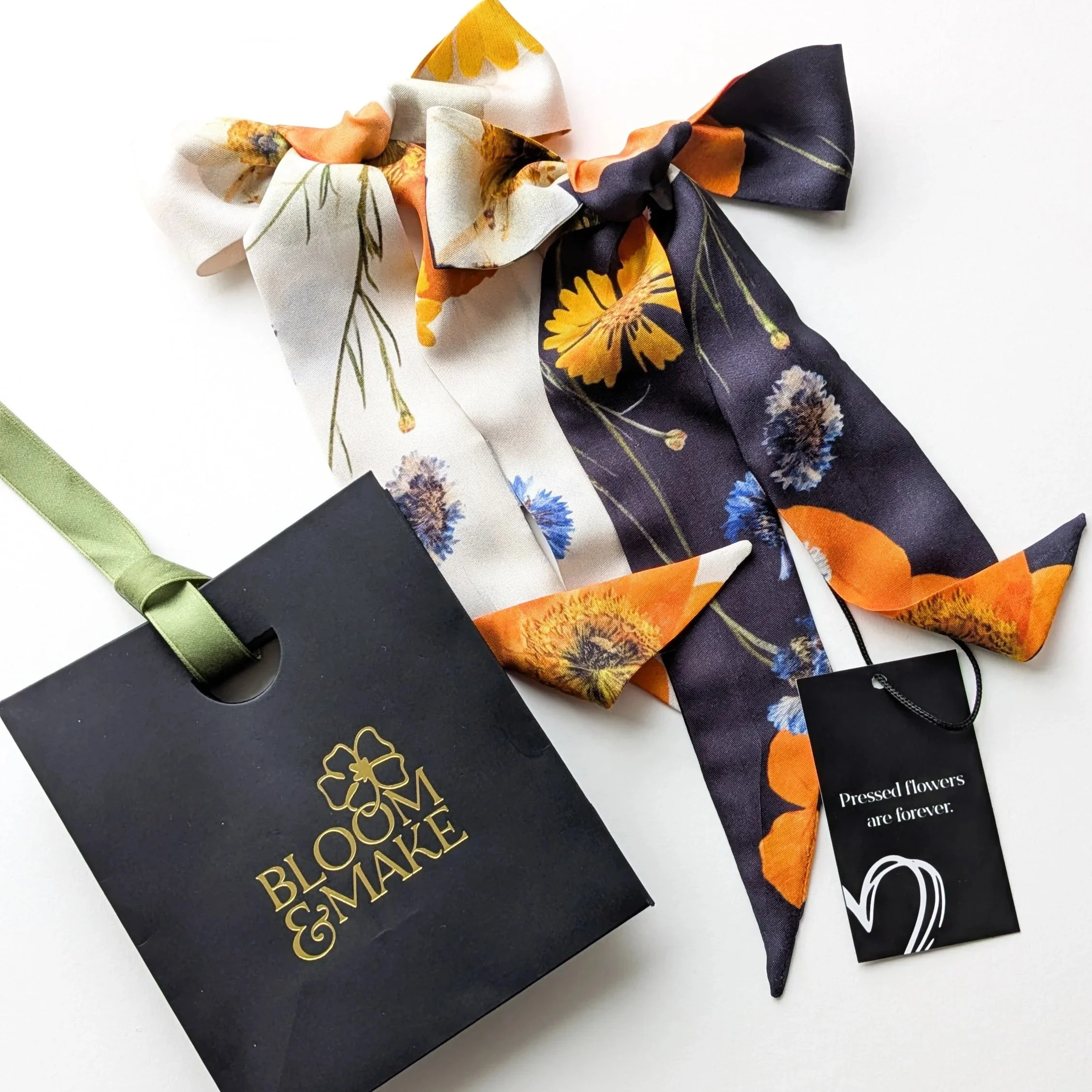 A black gift bag with a green ribbon handle, a gold flower logo, and the words 'BLOOM & MAKE'. Next to it, a floral-patterned silk scarf with orange, yellow, blue, and white flowers, includes a black tag with the phrase 'Pressed flowers are forever' attached by a black string.