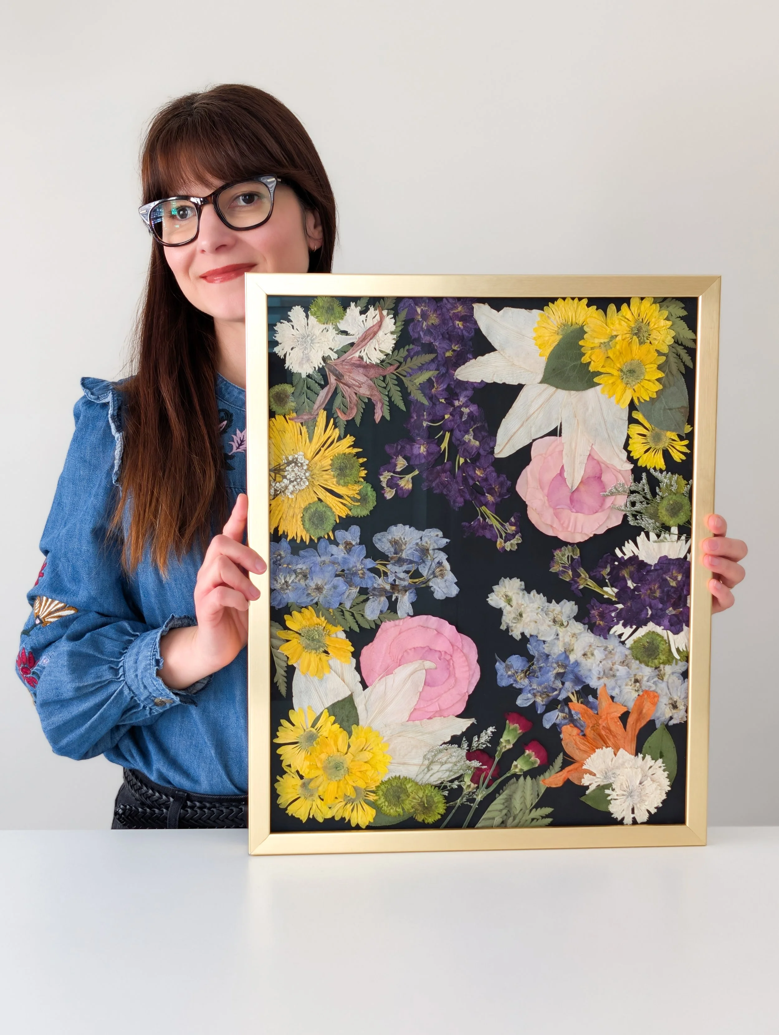 Woman with glasses holding a framed collection of pressed flowers.