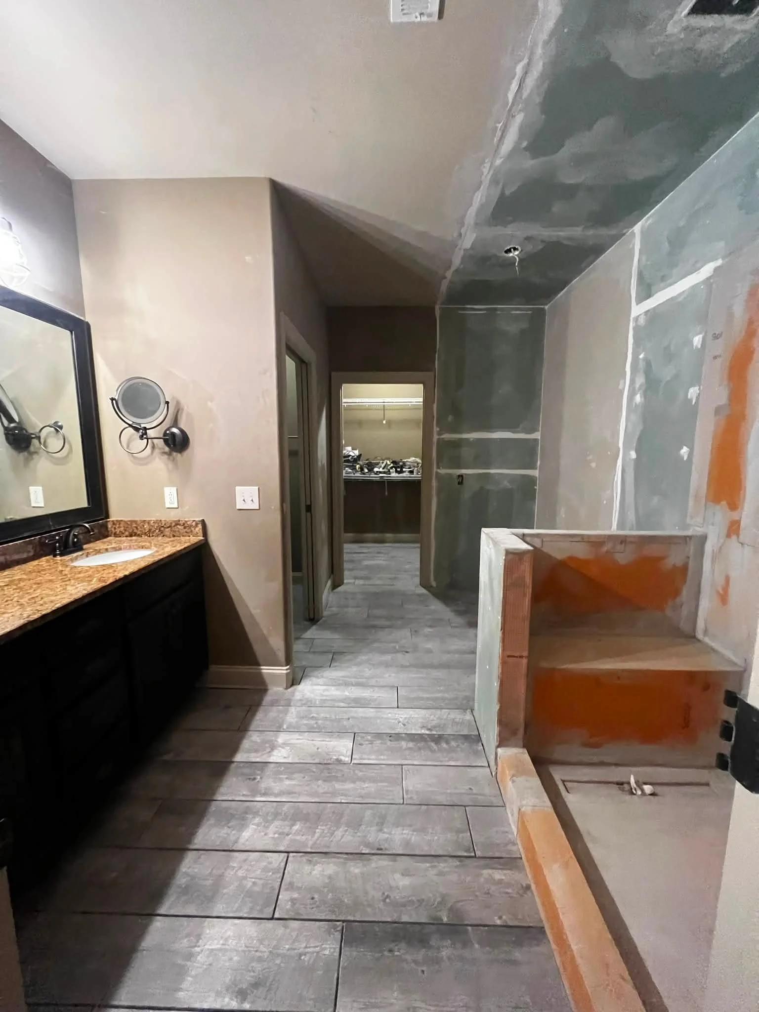 Bathroom under renovation with unfinished drywall, a granite countertop with a sink, wall-mounted mirror, and wood framing for a shower or bathtub.