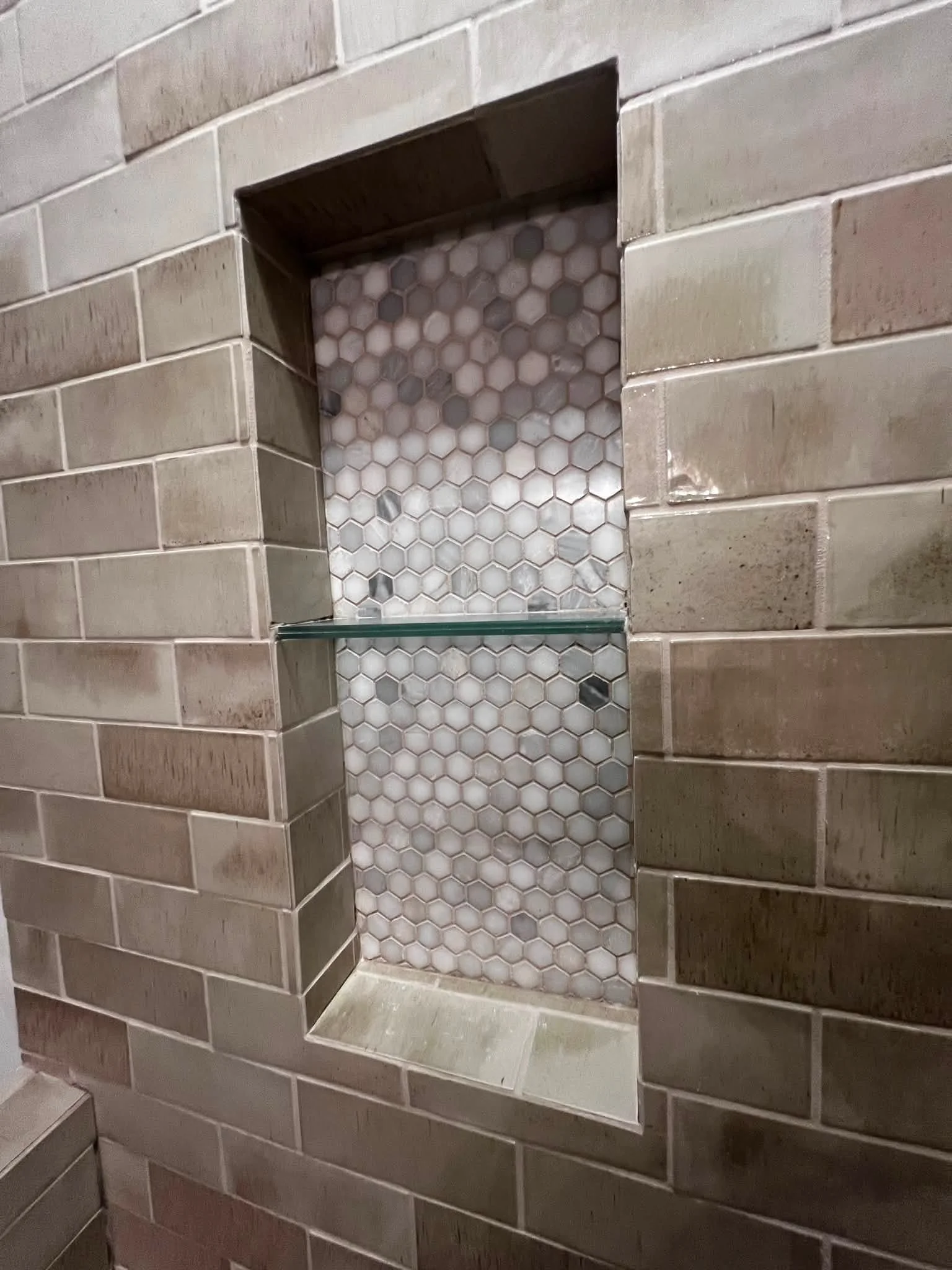 A close-up of a tiled wall with a small built-in shelf and a decorative tile inlay area. The wall has rectangular tiles in varying shades of beige and brown.