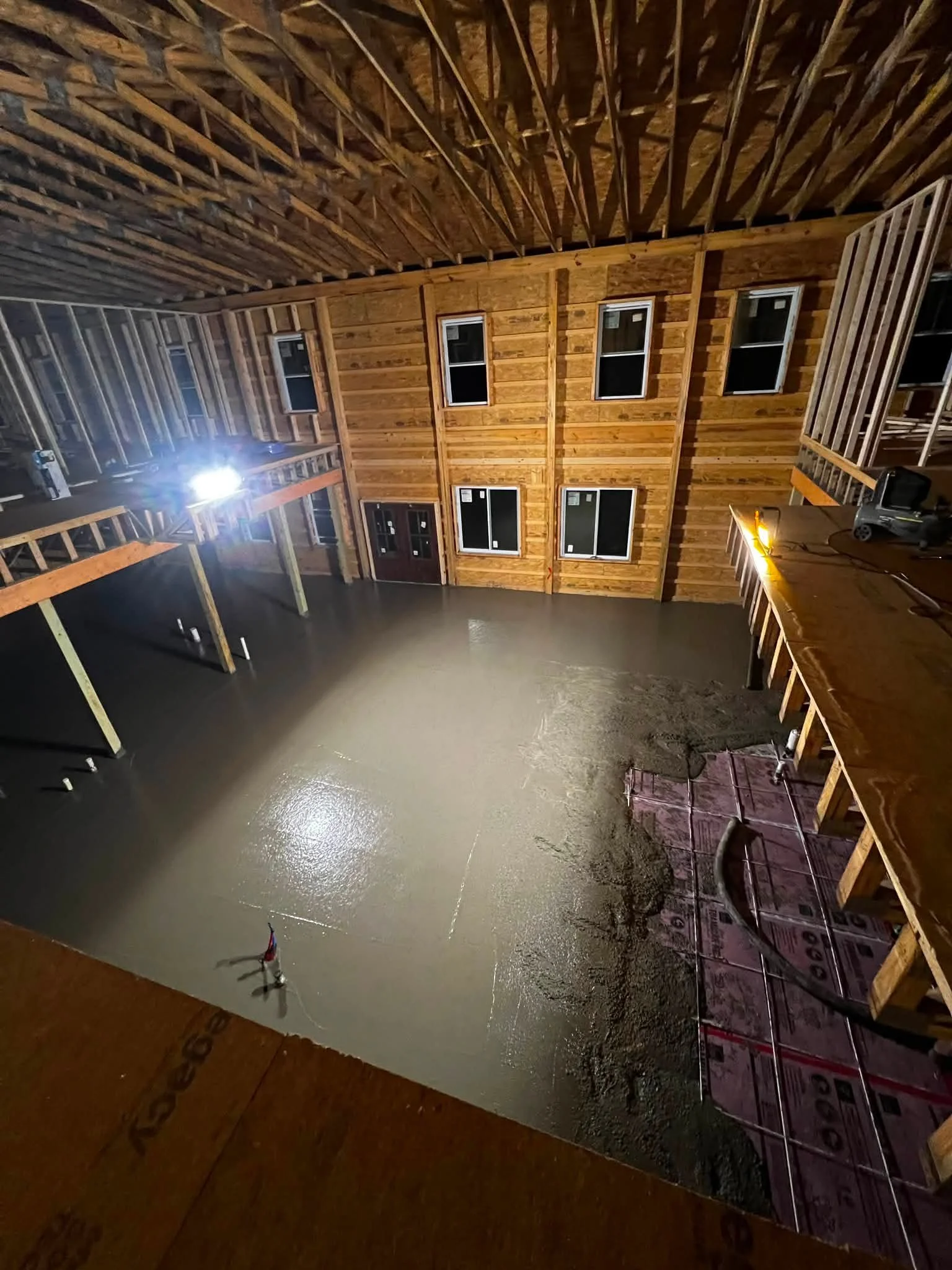 Interior view of a two-story unfinished house with exposed wood framing, windows, a staircase, and a polished concrete floor.