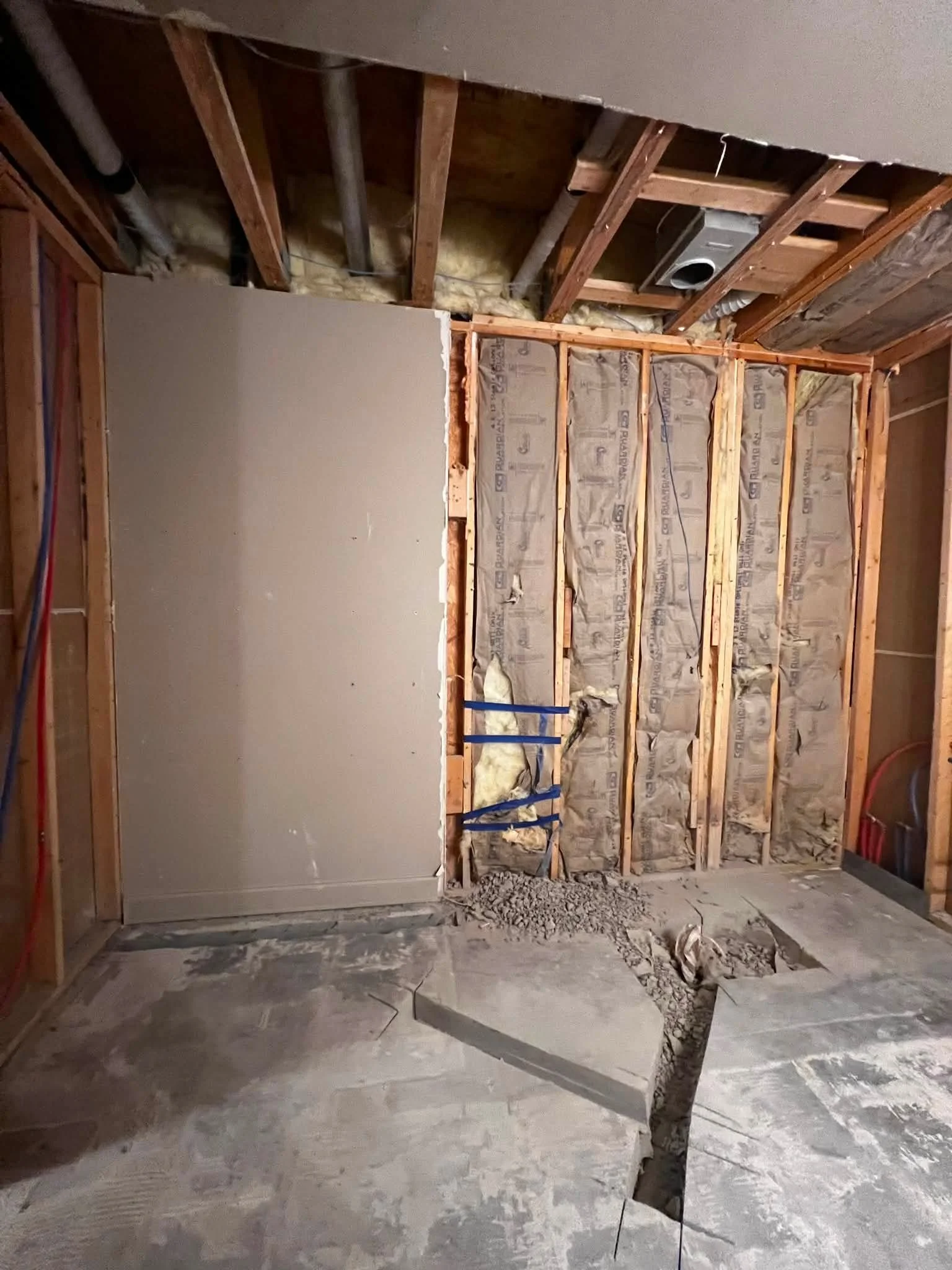 A partially constructed room with exposed wooden framing, insulation in the wall, some drywall installed, and a section of the floor dug out, possibly for plumbing or electrical work.