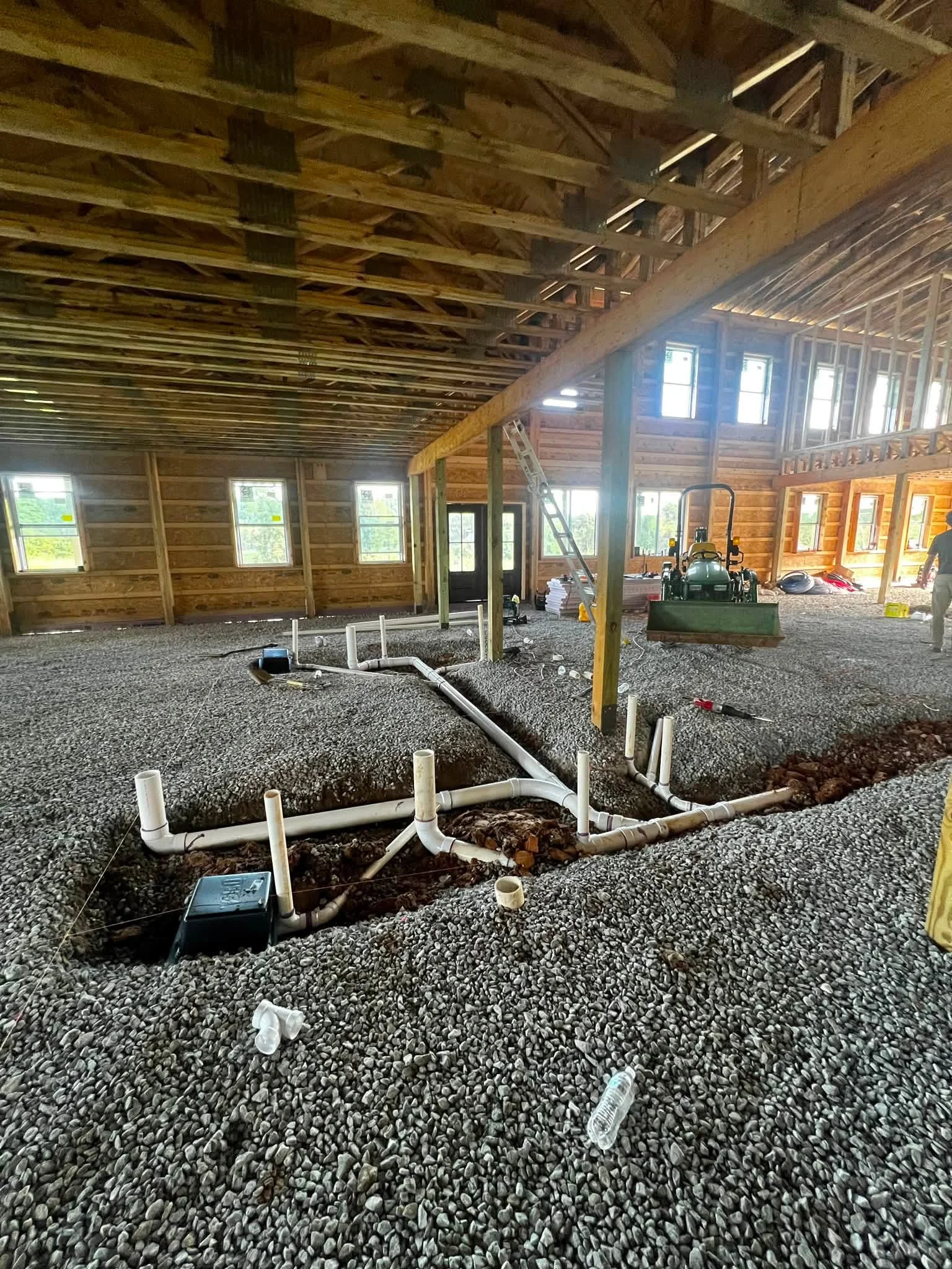 Interior of a house under construction with exposed wooden framing, multiple windows, and plumbing pipes on a gravel floor.