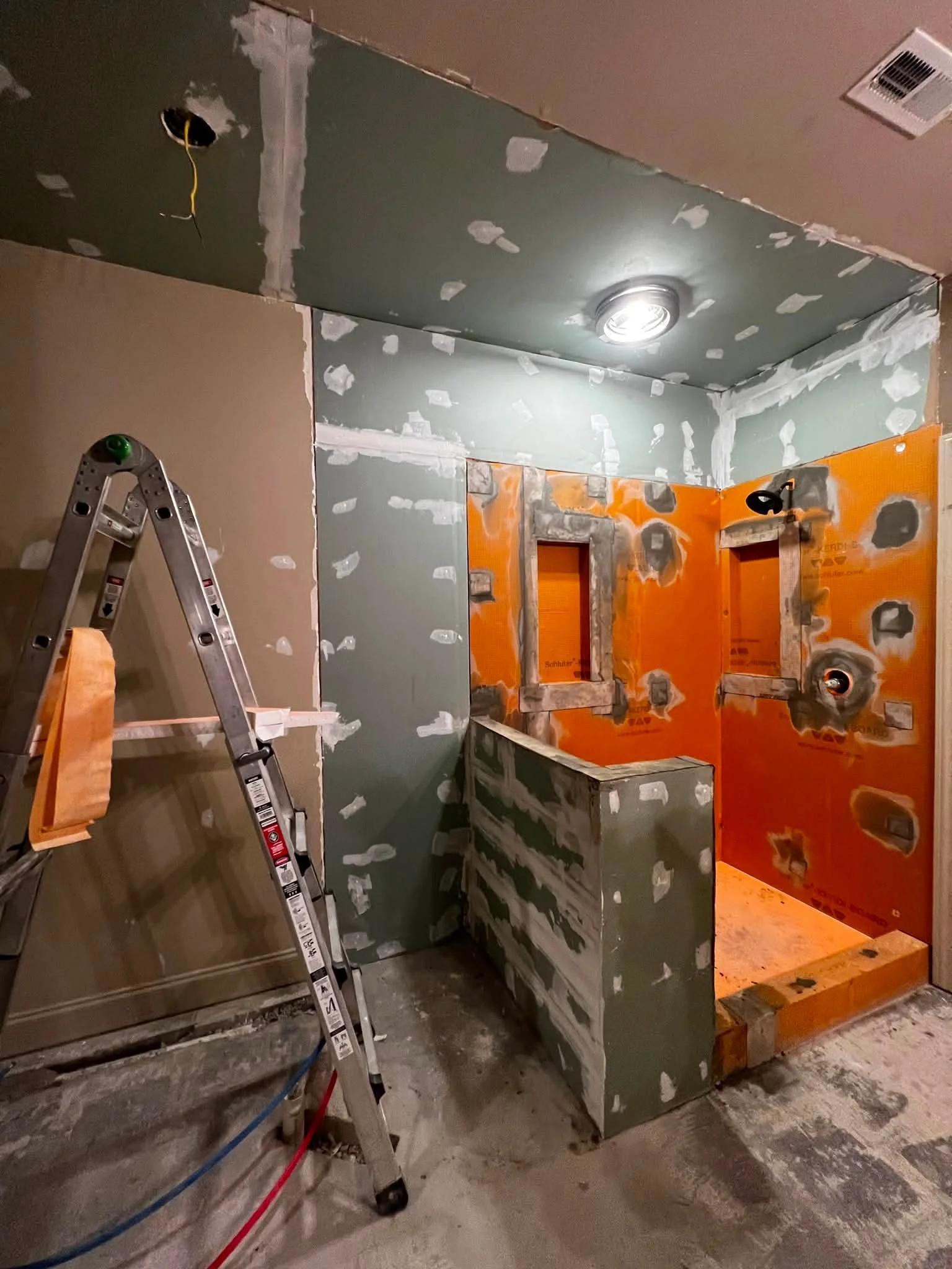 Bathroom under renovation with drywall installation, patching, and wiring visible, ladder and construction materials present.