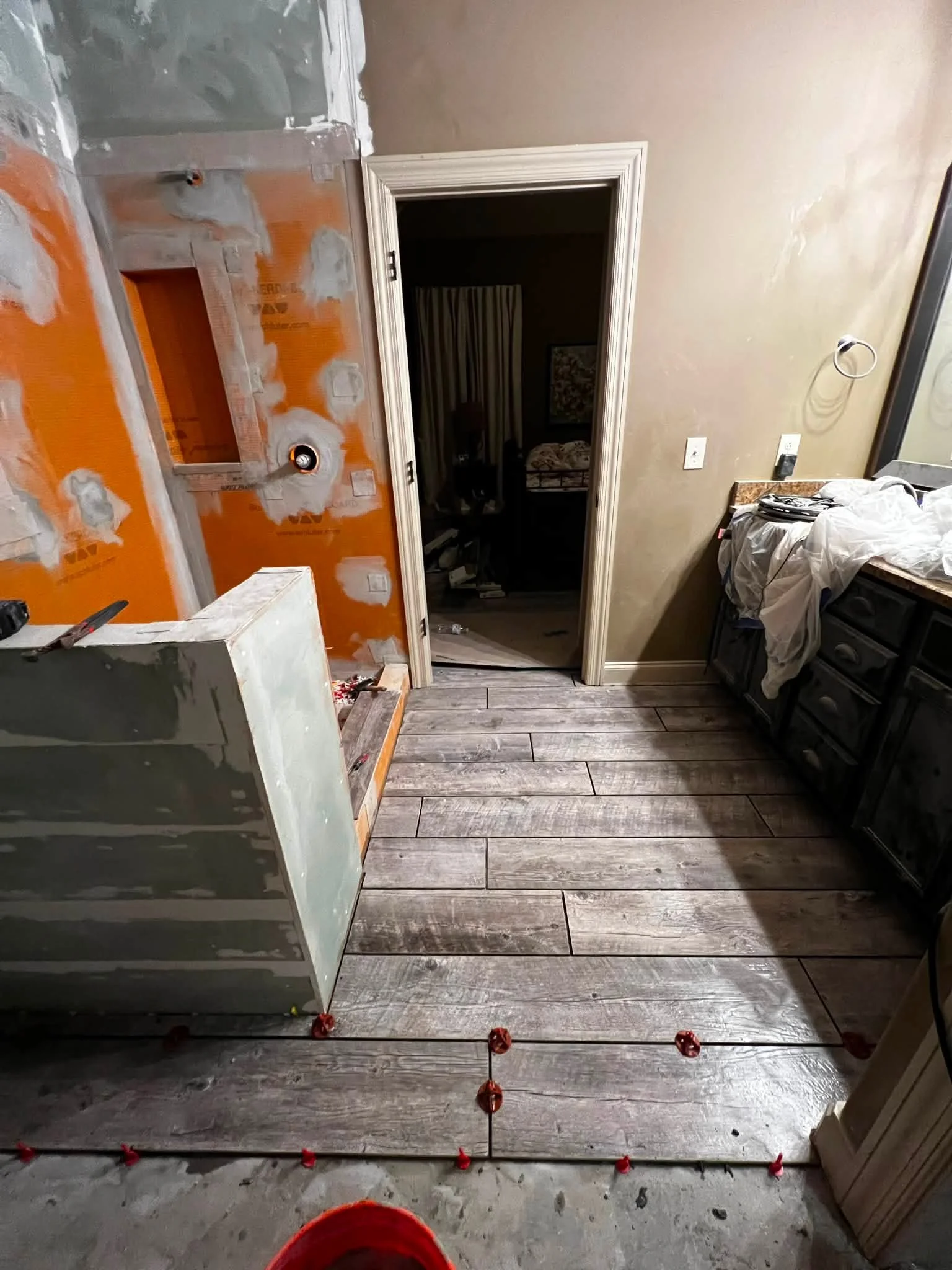 Interior of a room under renovation with partially painted orange walls, an open doorway leading to a dark bedroom, and construction tools and materials scattered around including a level and some red markers on the floor.