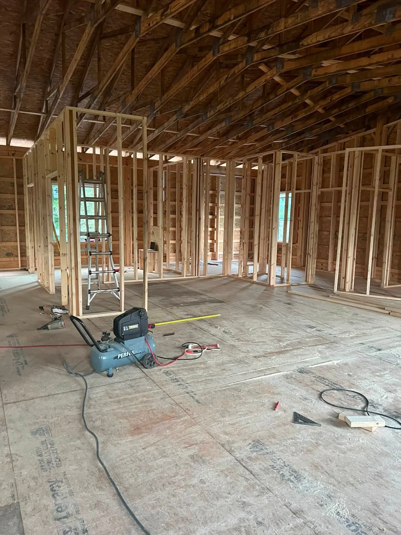 Interior view of a house under construction with exposed wooden framing, a ladder, construction tools, and equipment on the unfinished wooden floor.