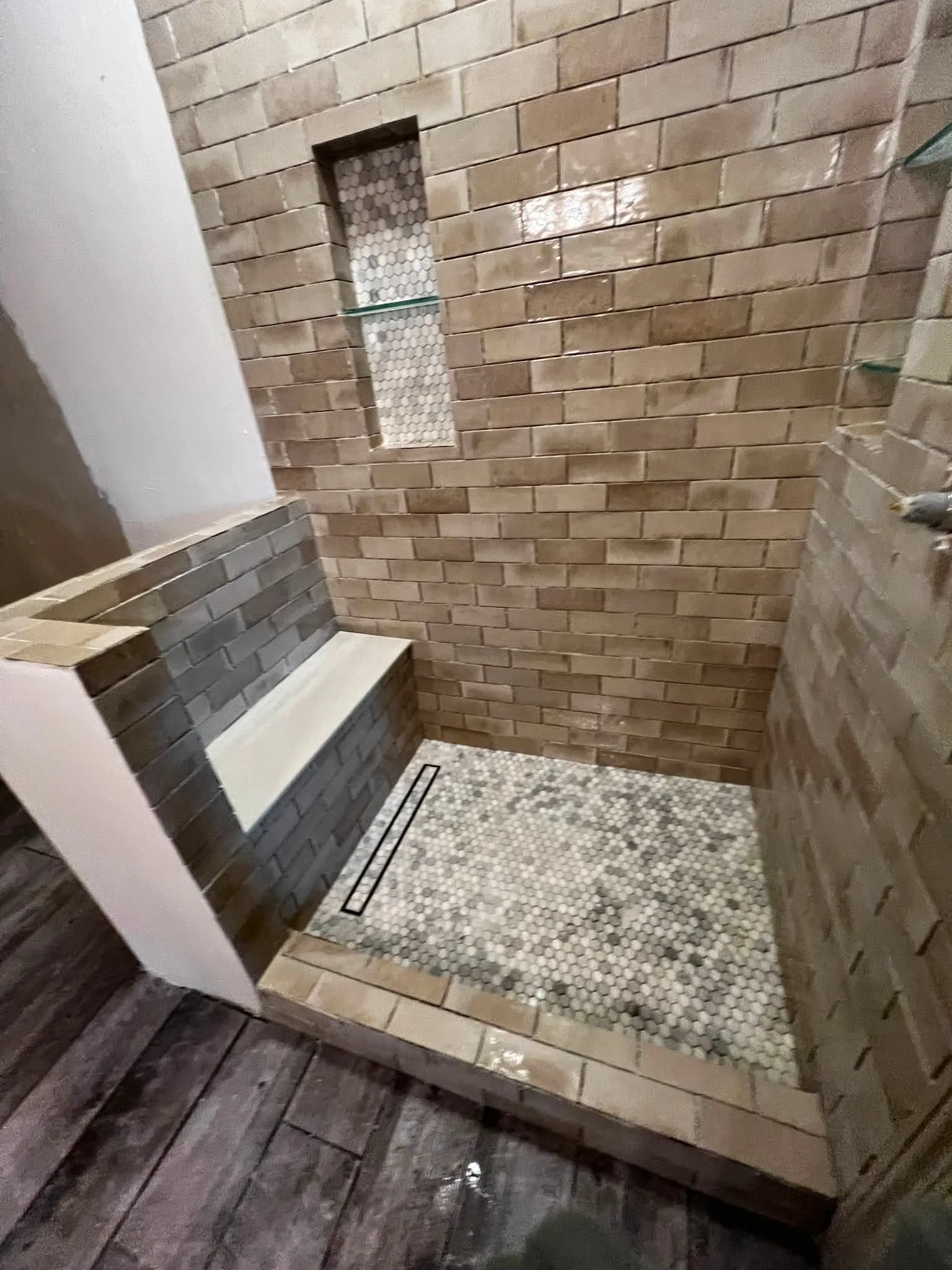 Interior of a bathroom or shower area with brick tile walls, mosaic tile flooring, a bench, and a built-in niche with glass shelf.