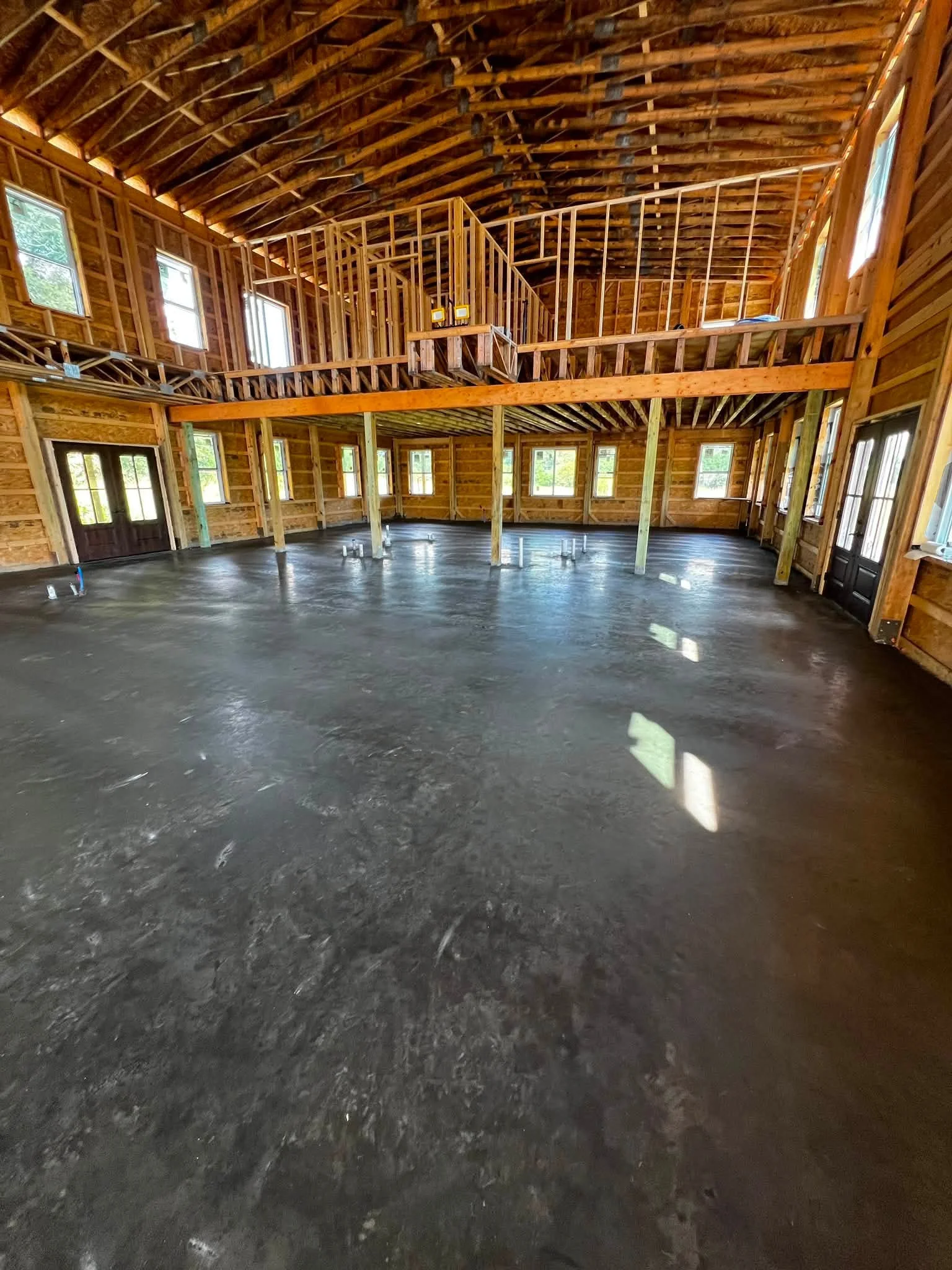 Interior of a house under construction with wooden framing, high ceilings, multiple windows, and a concrete floor.