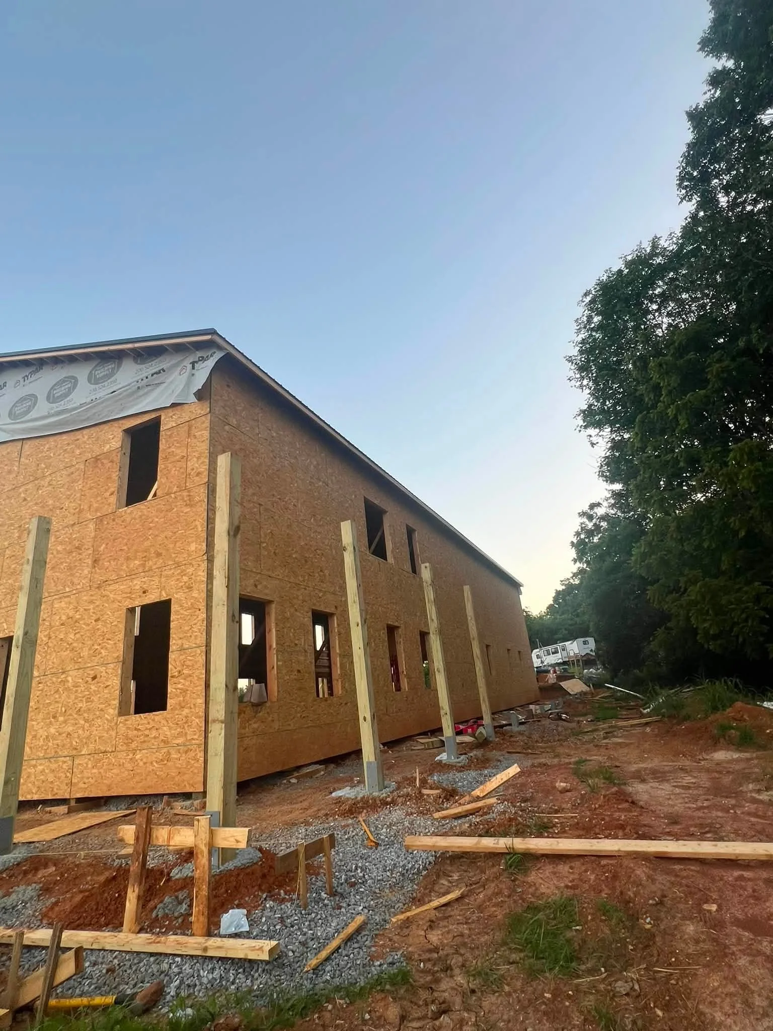 A house under construction with plywood walls and window openings, surrounded by construction materials and wooden supports, with trees and a clear sky in the background.