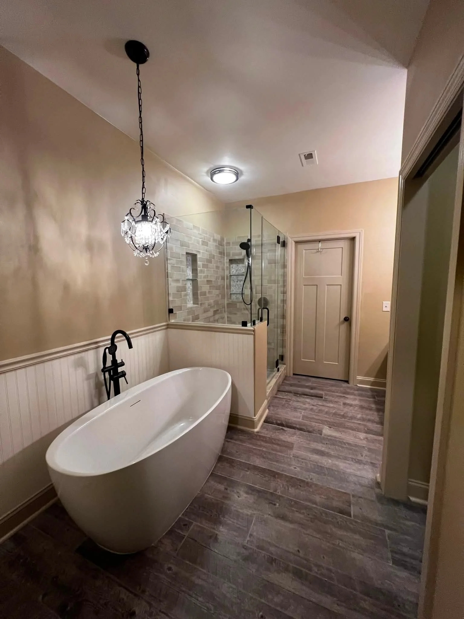 A modern bathroom featuring a freestanding bathtub, a glass-enclosed shower with brick-style tile, a chandelier, and wood-look flooring.