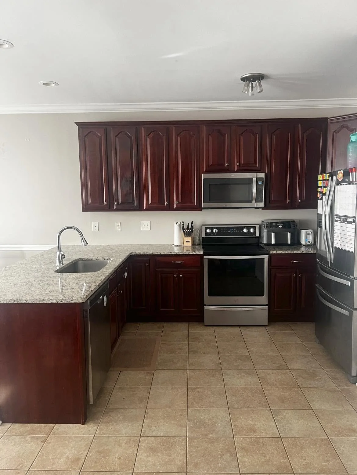Kitchen with dark wooden cabinets, granite countertops, stainless steel appliances including microwave, oven, toaster, and refrigerator, tiled floor, and a kitchen sink with a faucet.