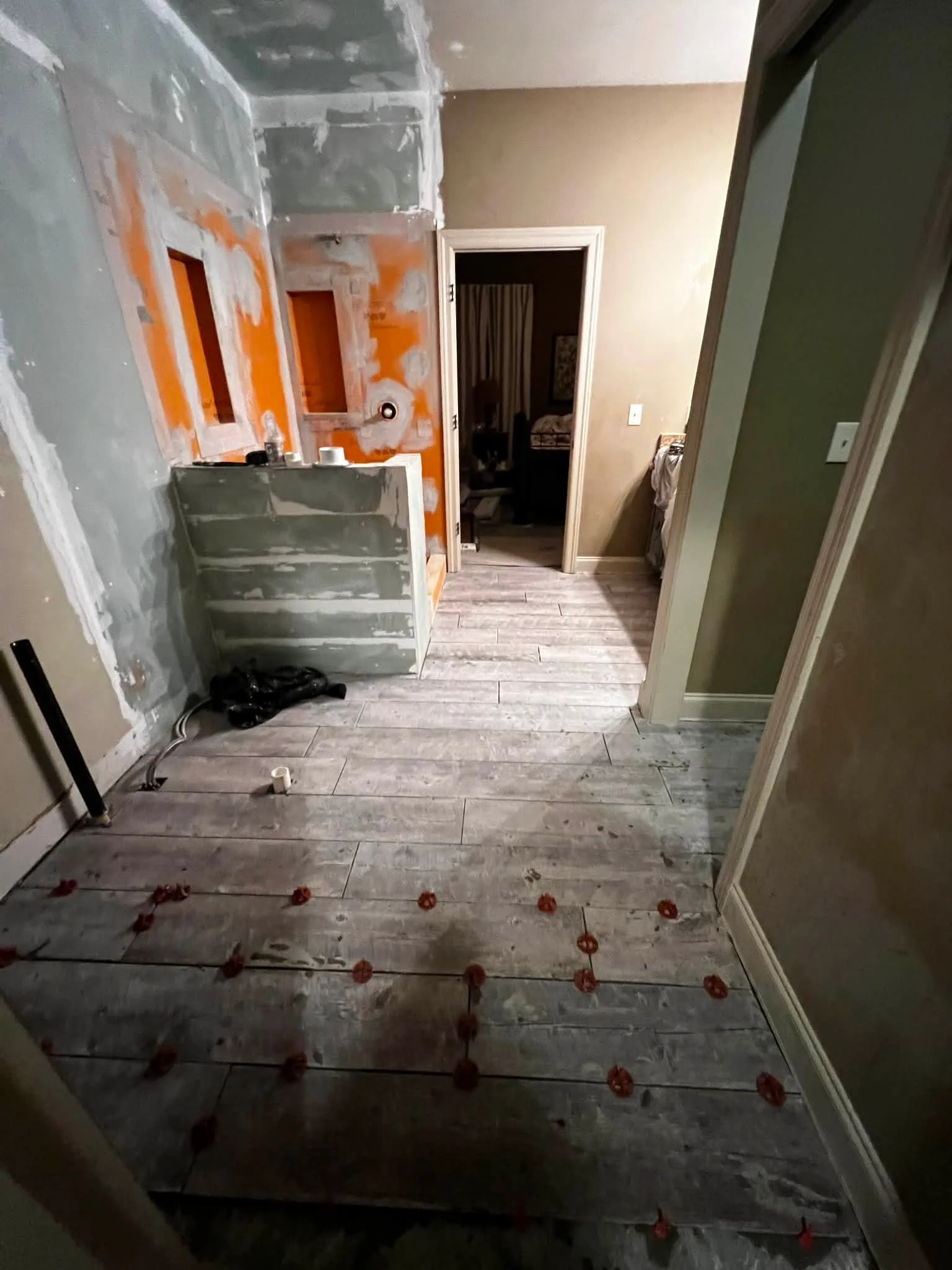Room under renovation with unfinished drywall, construction materials, and wooden floor. Tape measures or tools are visible, and safety markers or small orange decorations are lined along the floor.