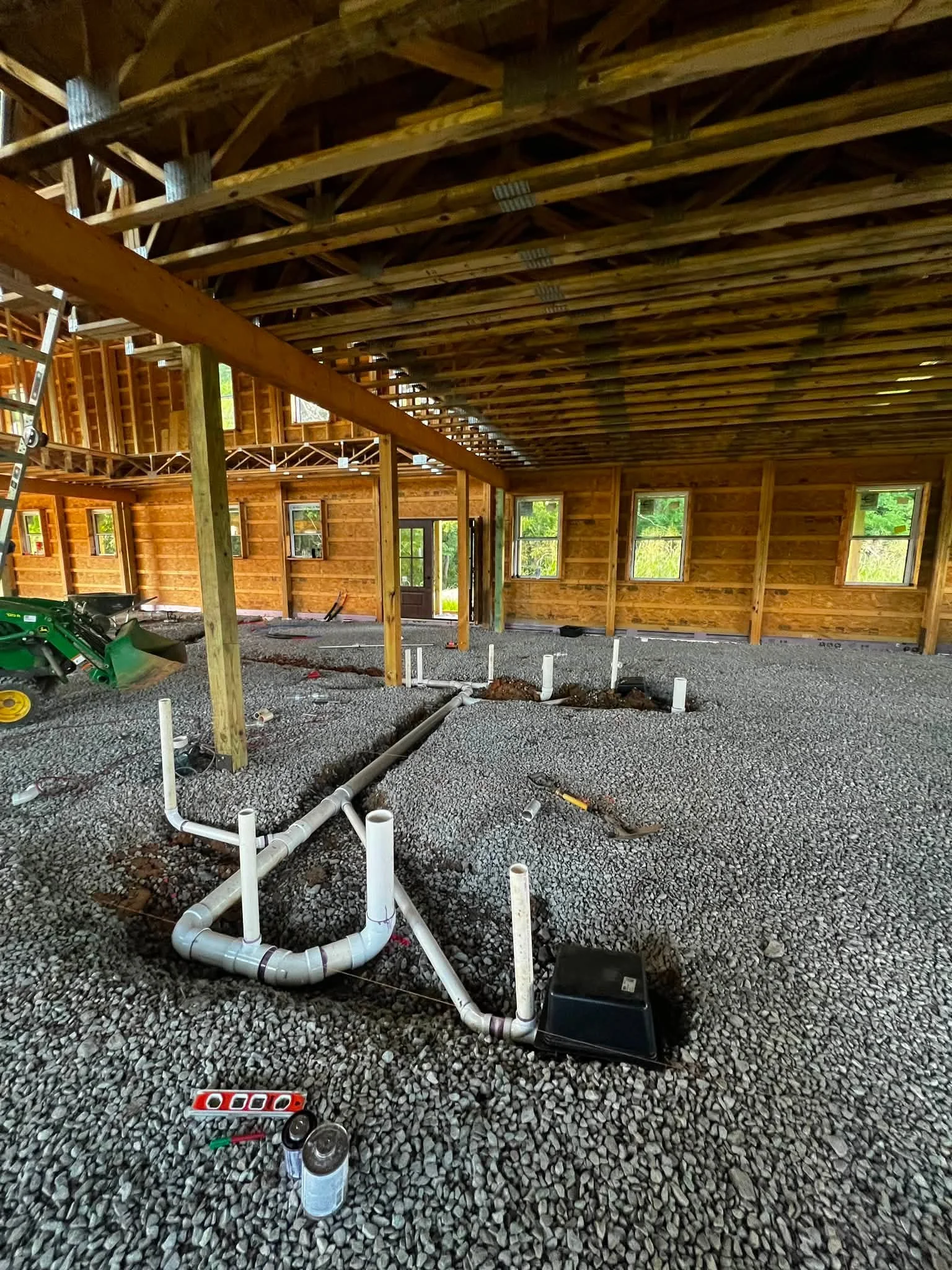 Interior of a building under construction with exposed wooden beams and framed windows, showing plumbing pipes and construction tools on a gravel floor.