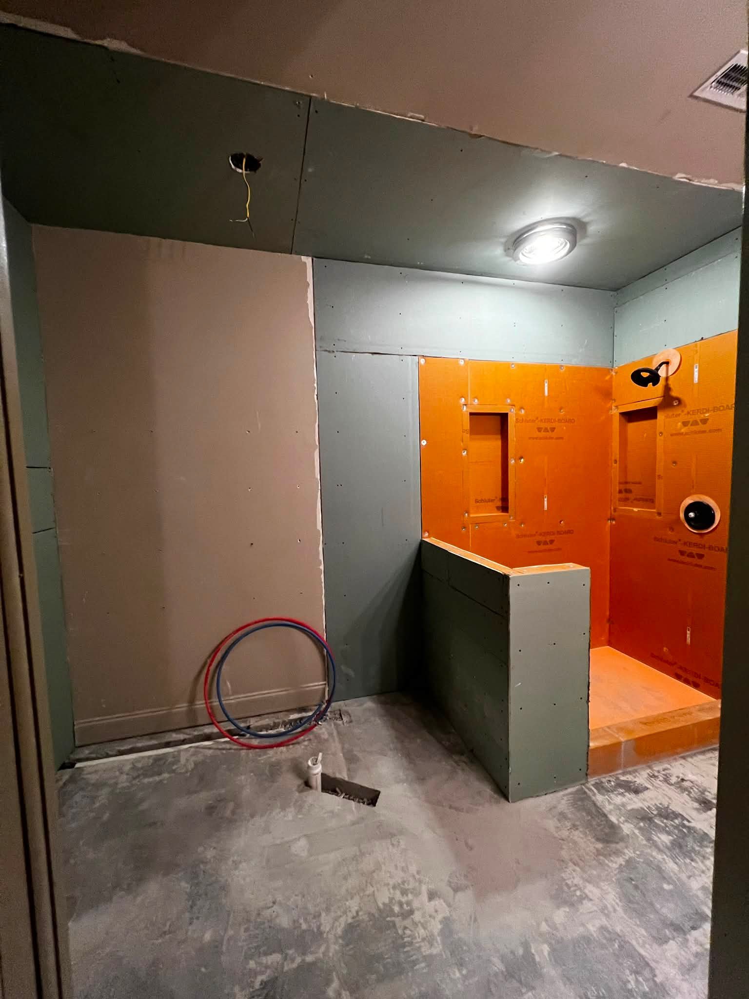 Interior of a room under construction with partially installed drywall; orange soundproofing panels on the right wall, exposed wires hanging from the ceiling, a ceiling-mounted light fixture, and plumbing pipes on the floor.