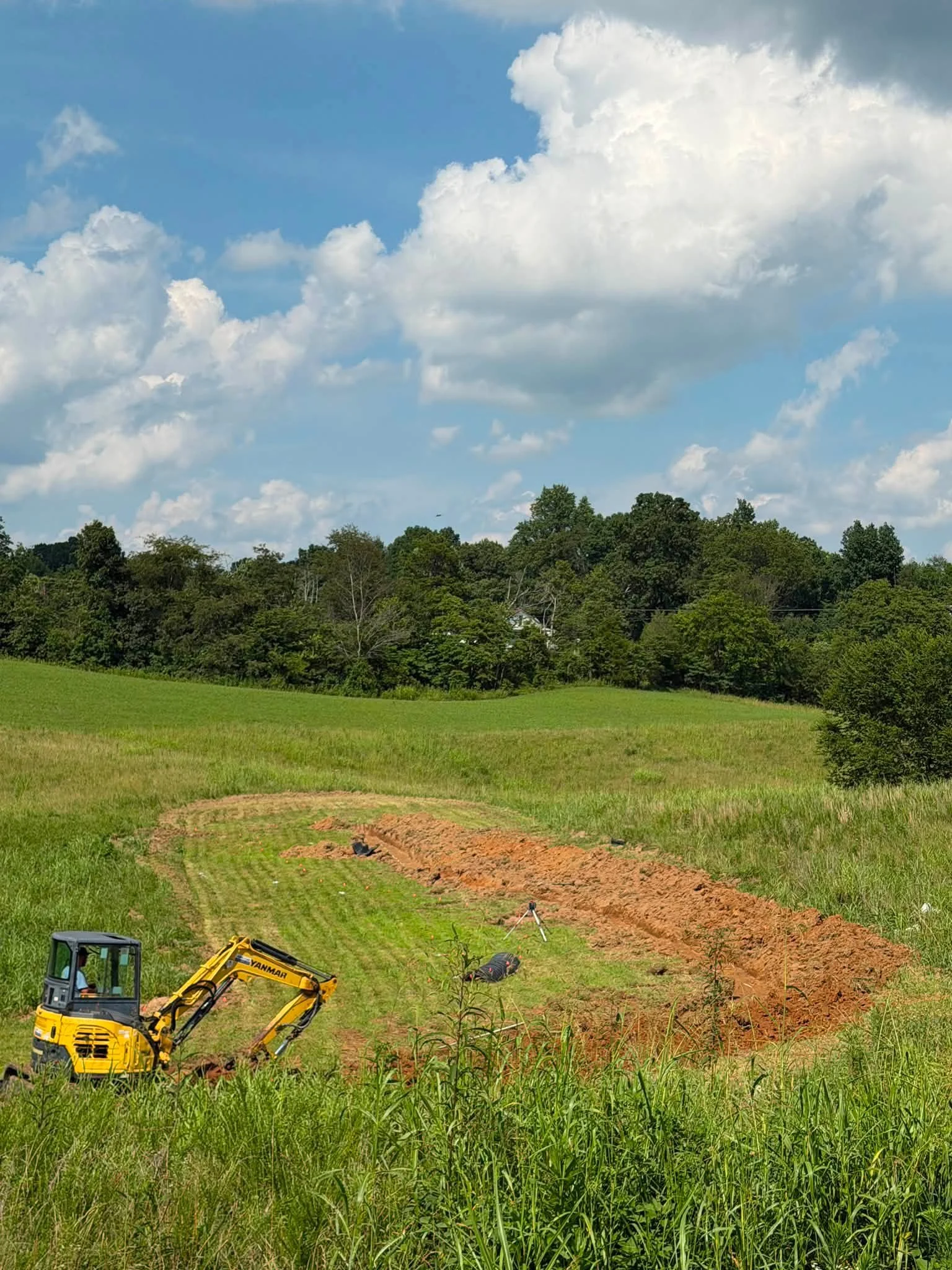 A construction site in a grassy field with a yellow mini excavator, a tripod, and some tools, with a backdrop of trees and a blue sky with white clouds.