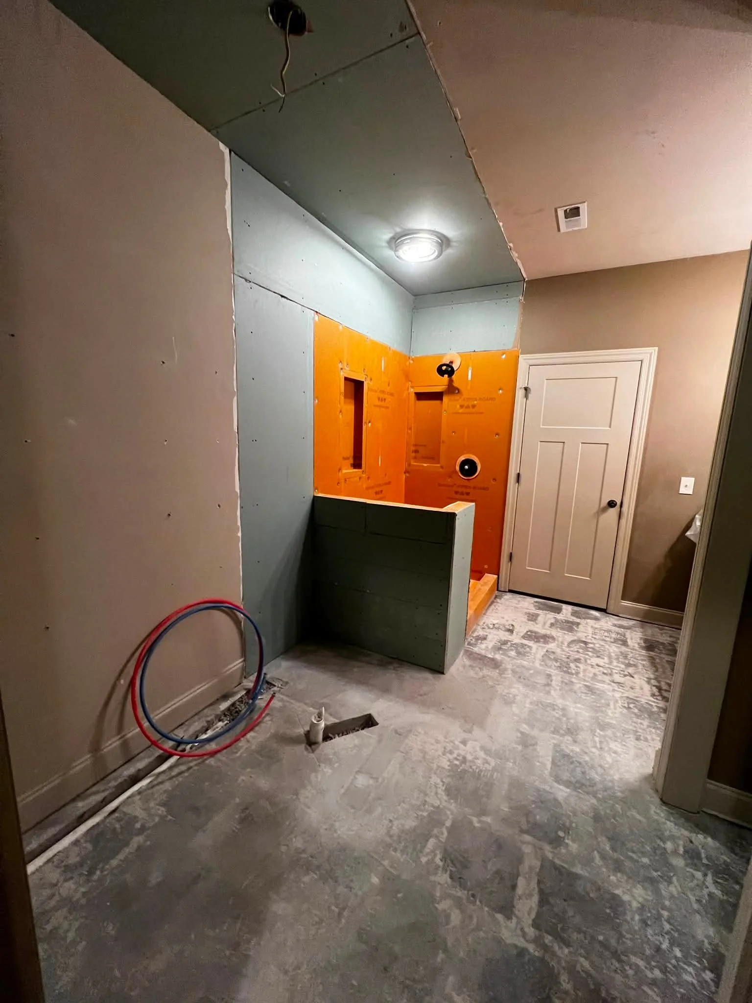 A room under renovation with unfinished walls, exposed wiring, and plumbing, with a door on the right side and a ceiling light fixture.