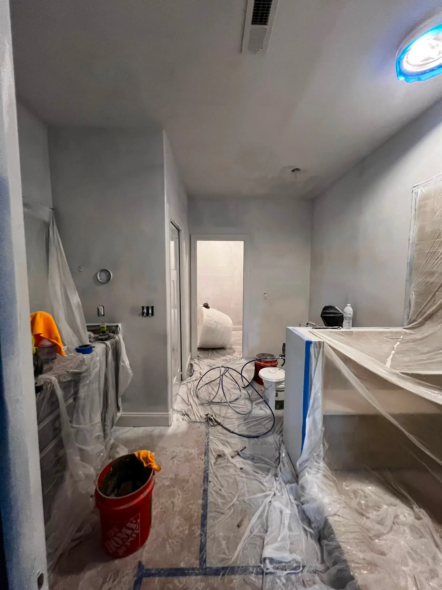 A room under renovation with plastic covers, tape, and painting tools scattered around. The ceiling has a vent, and the walls are painted light gray. There are some paint supplies and a water bottle on the counter.