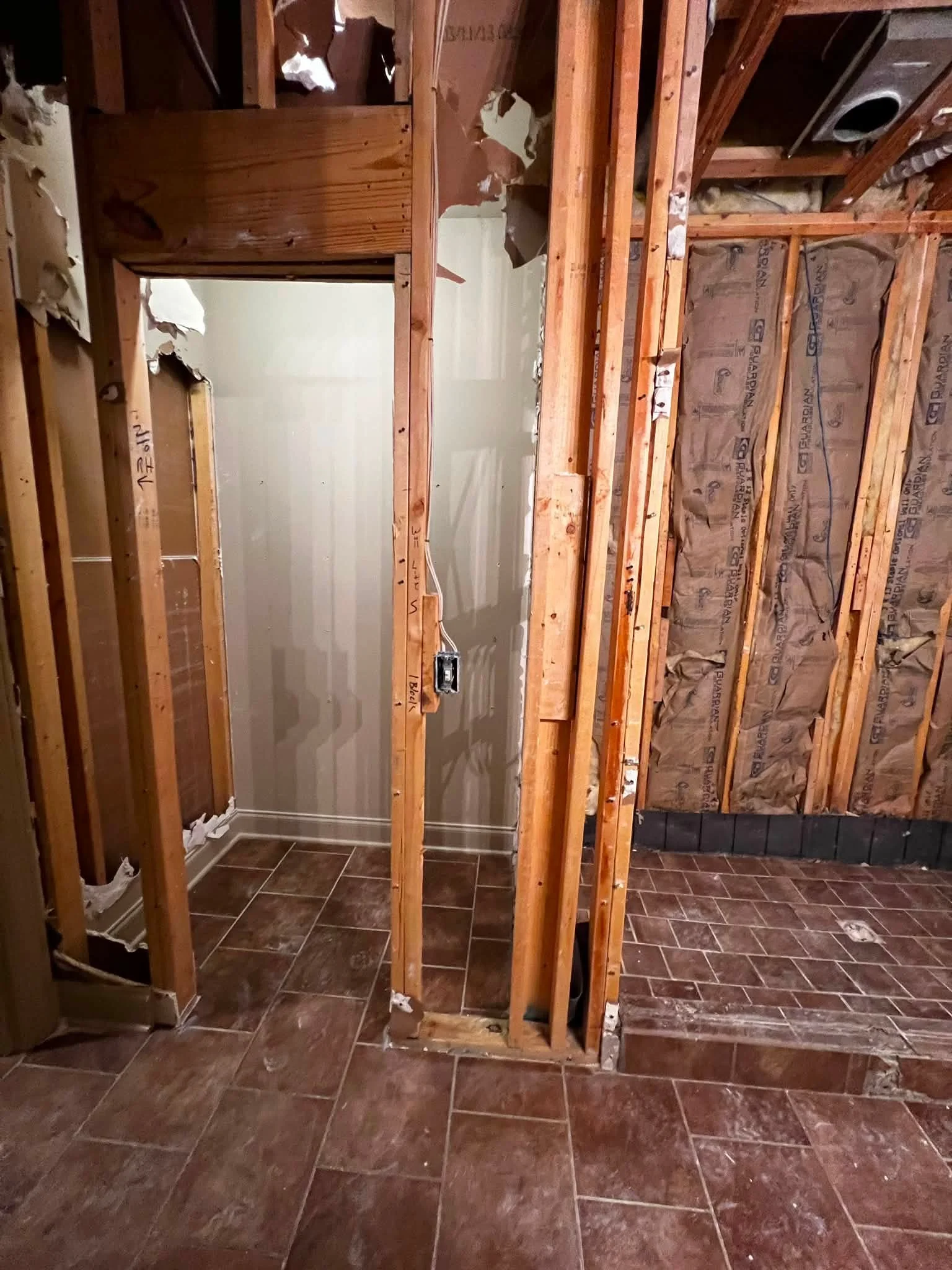 Partially demolished interior wall with exposed wooden framework and insulation, brown tiled floor, and a doorway leading to another room.