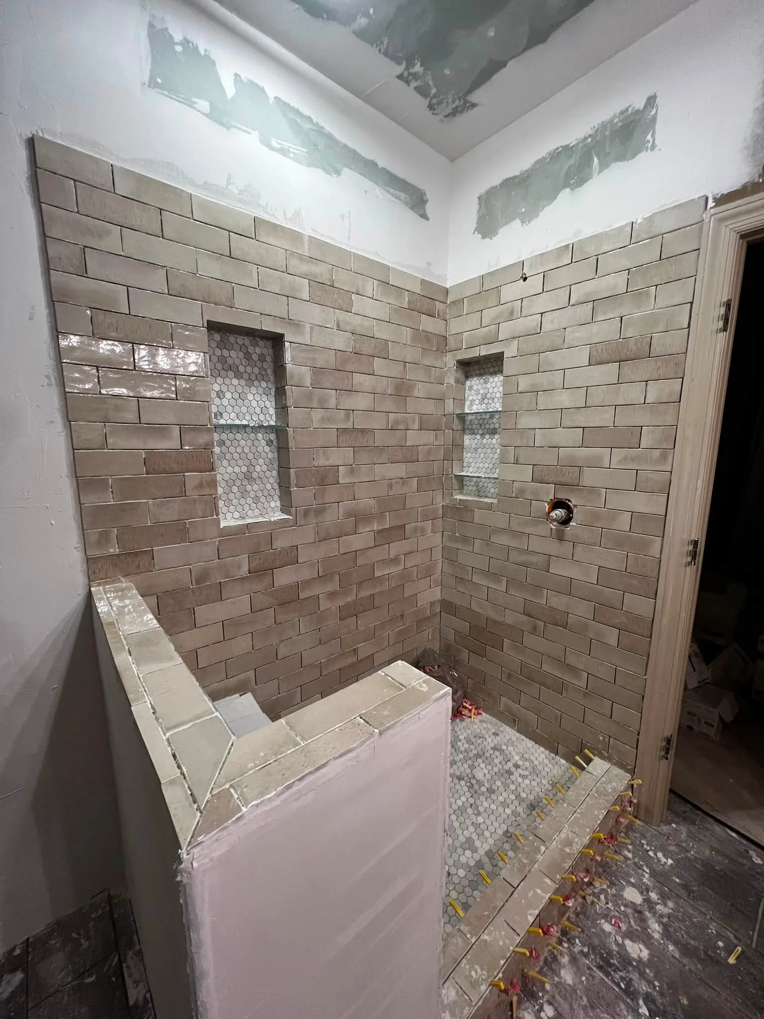 A bathroom under renovation with beige subway tiles on the walls and hexagon tiles on the floor and in the wall niches. The walls are partially tiled, and there is drywall with unpainted patchwork on the ceiling. Some construction materials and tools