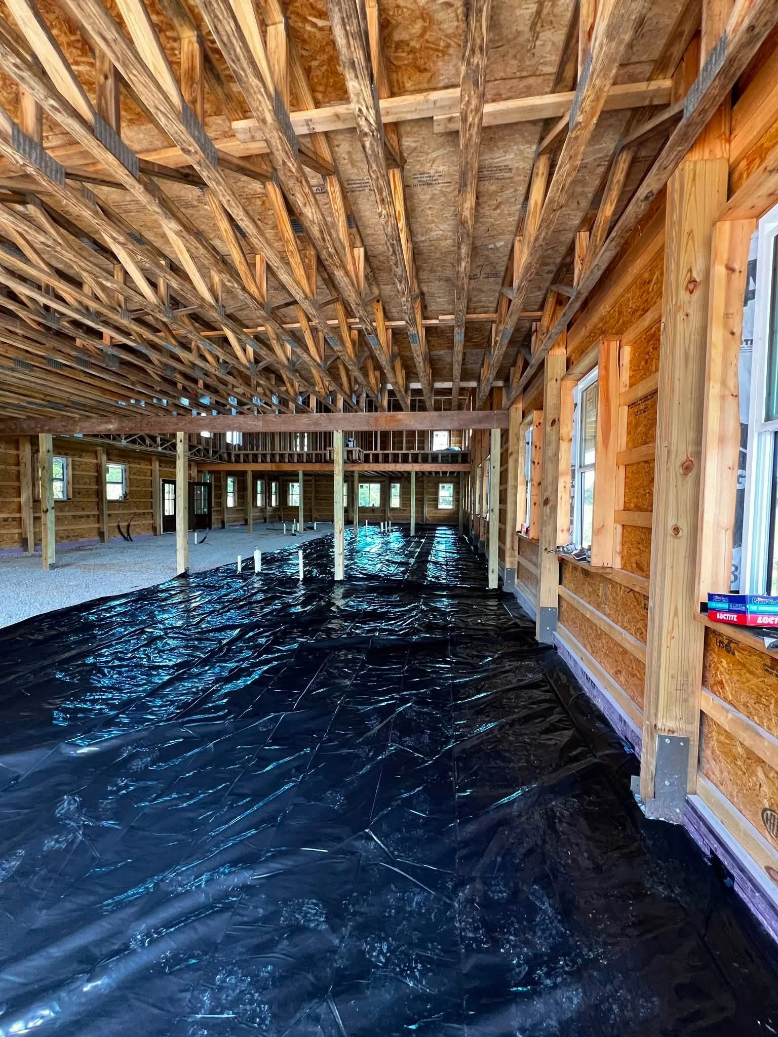 The interior of a house under construction with exposed wooden framework, studs, and a black plastic vapor barrier on the floor.