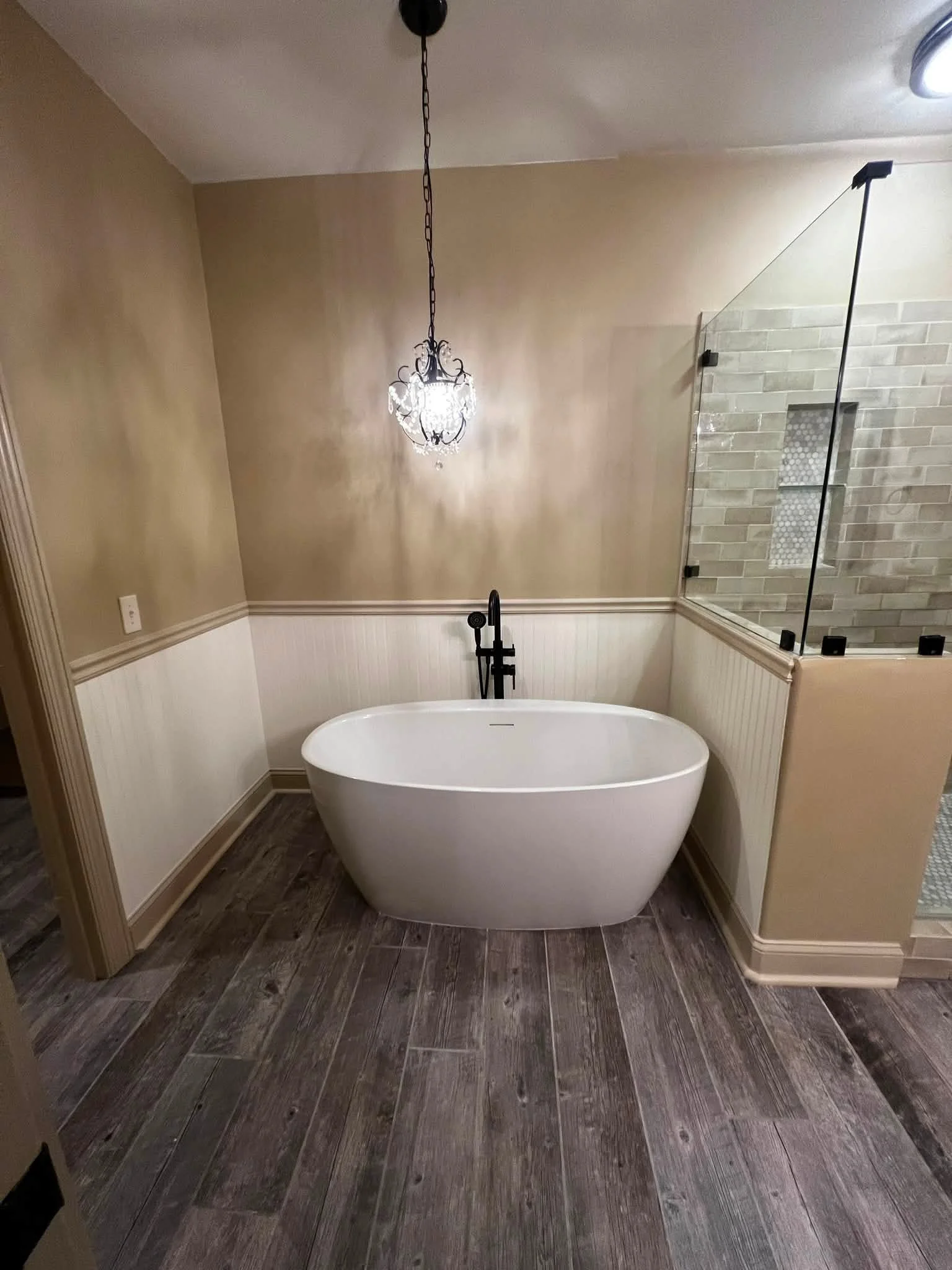 Bathroom with dark wood flooring, beige walls with white paneling, a freestanding white bathtub, a chandelier, and a walk-in shower with glass enclosure and tiled wall.