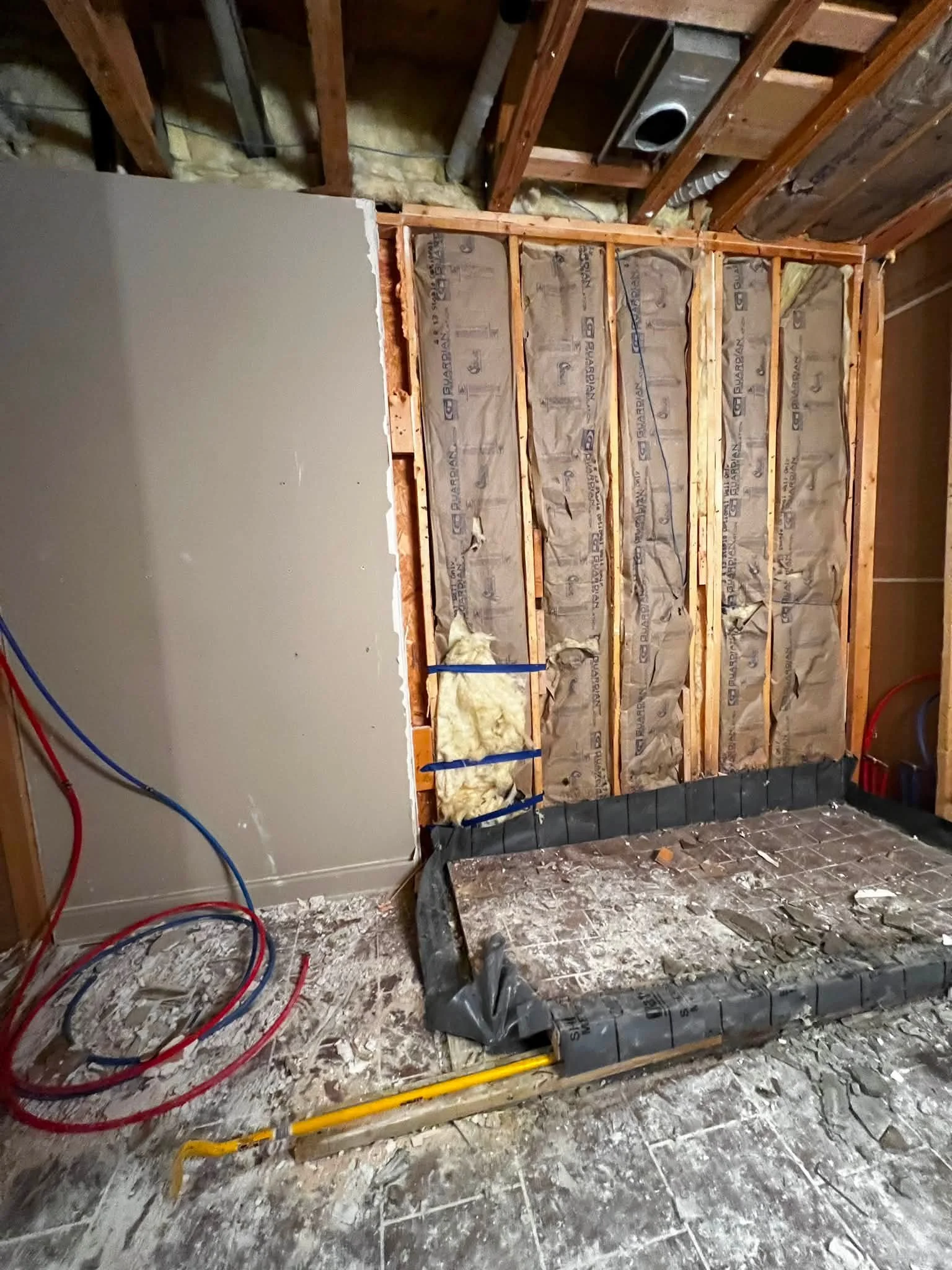 Interior of a room under construction showing exposed studs, insulation, and electrical wiring, with a partially installed wall and construction debris on the floor.
